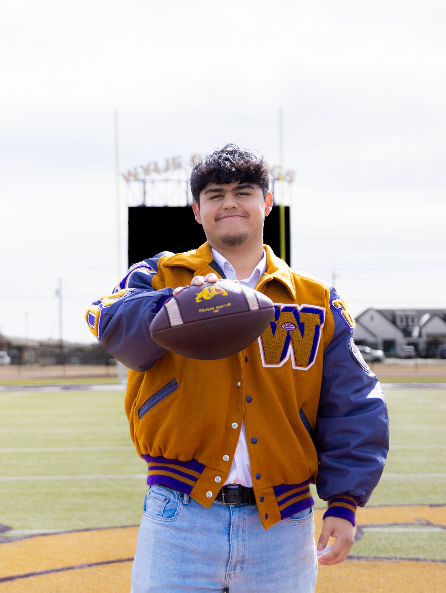 Young man in Wylie Bulldogs letterman jacket holding football on school field, pointing at camera.