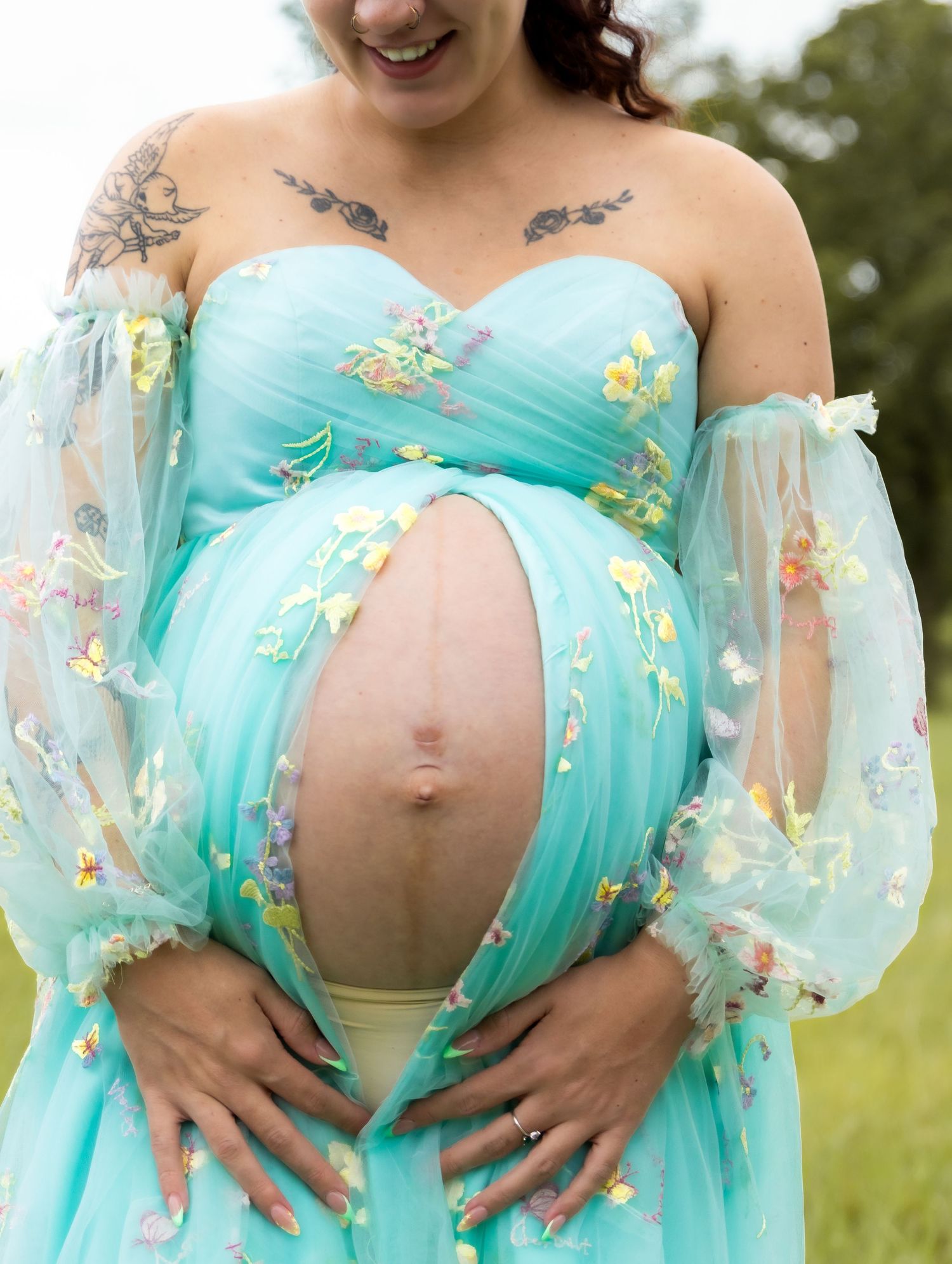Pregnant woman in teal floral gown with tattoos cradling bare baby bump outdoors.
