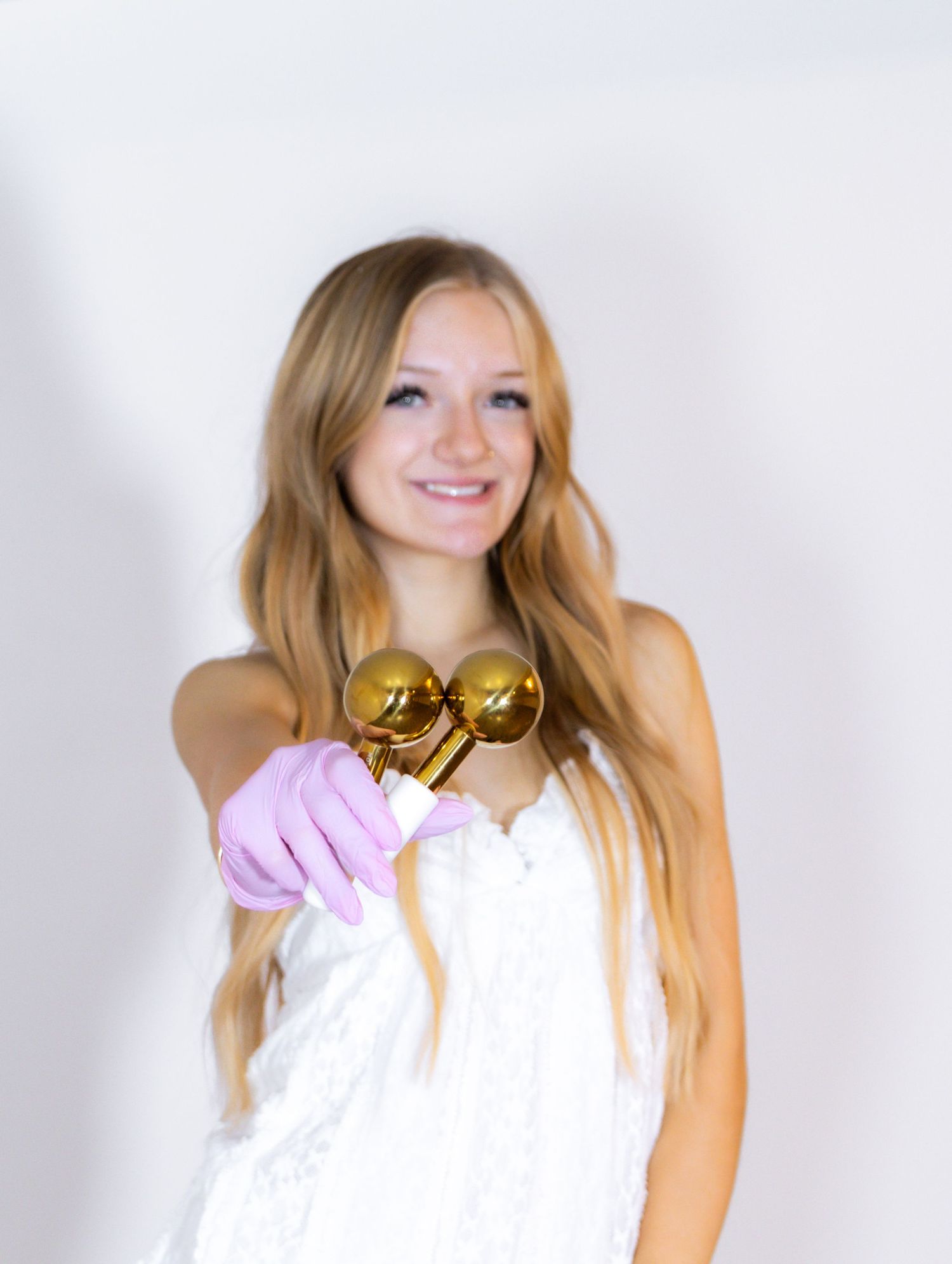 Smiling blonde girl in white dress holding gold maracas with pink glove against white background.