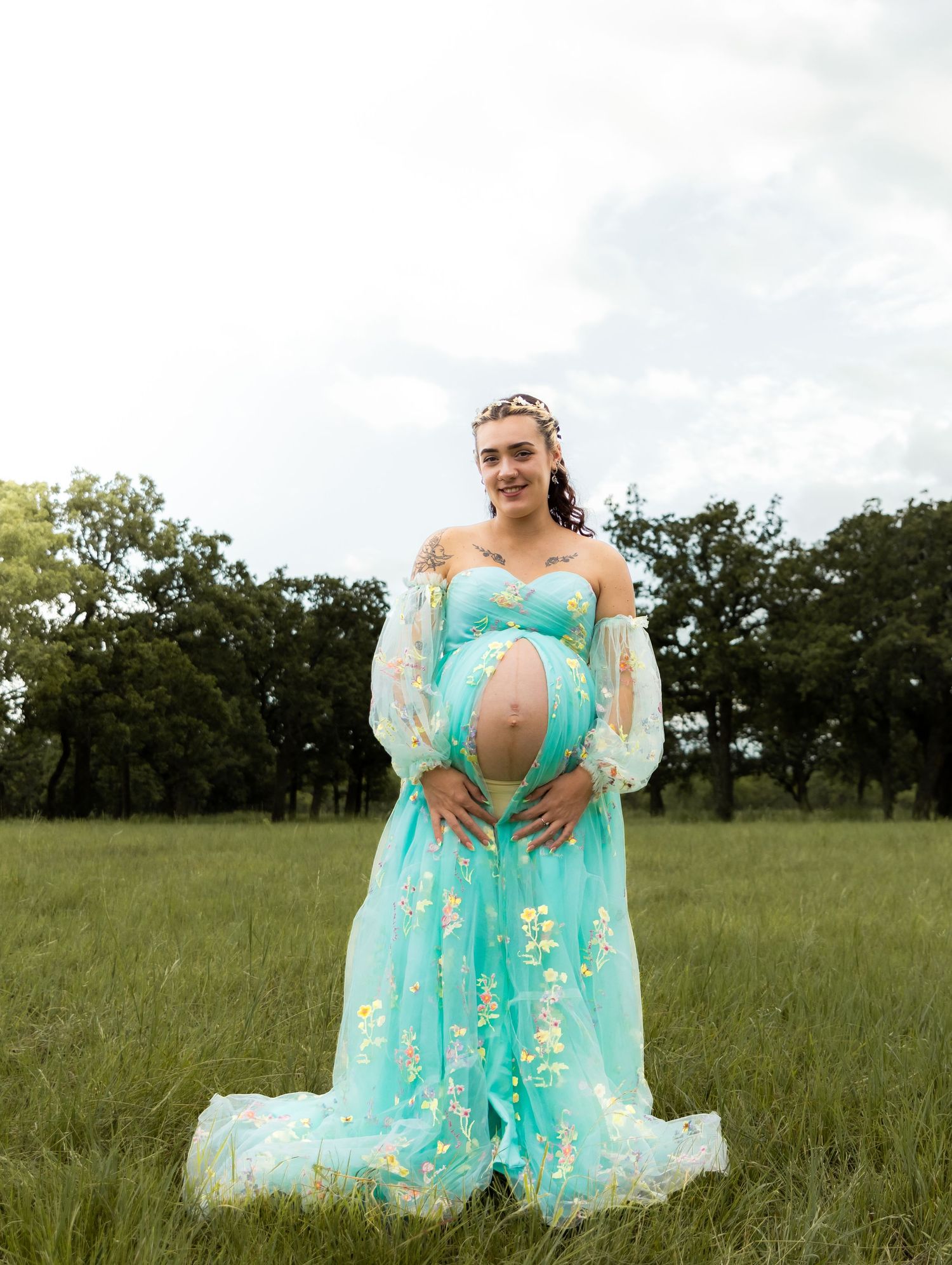 Pregnant woman in teal floral maternity gown cradling baby bump in lush green field.