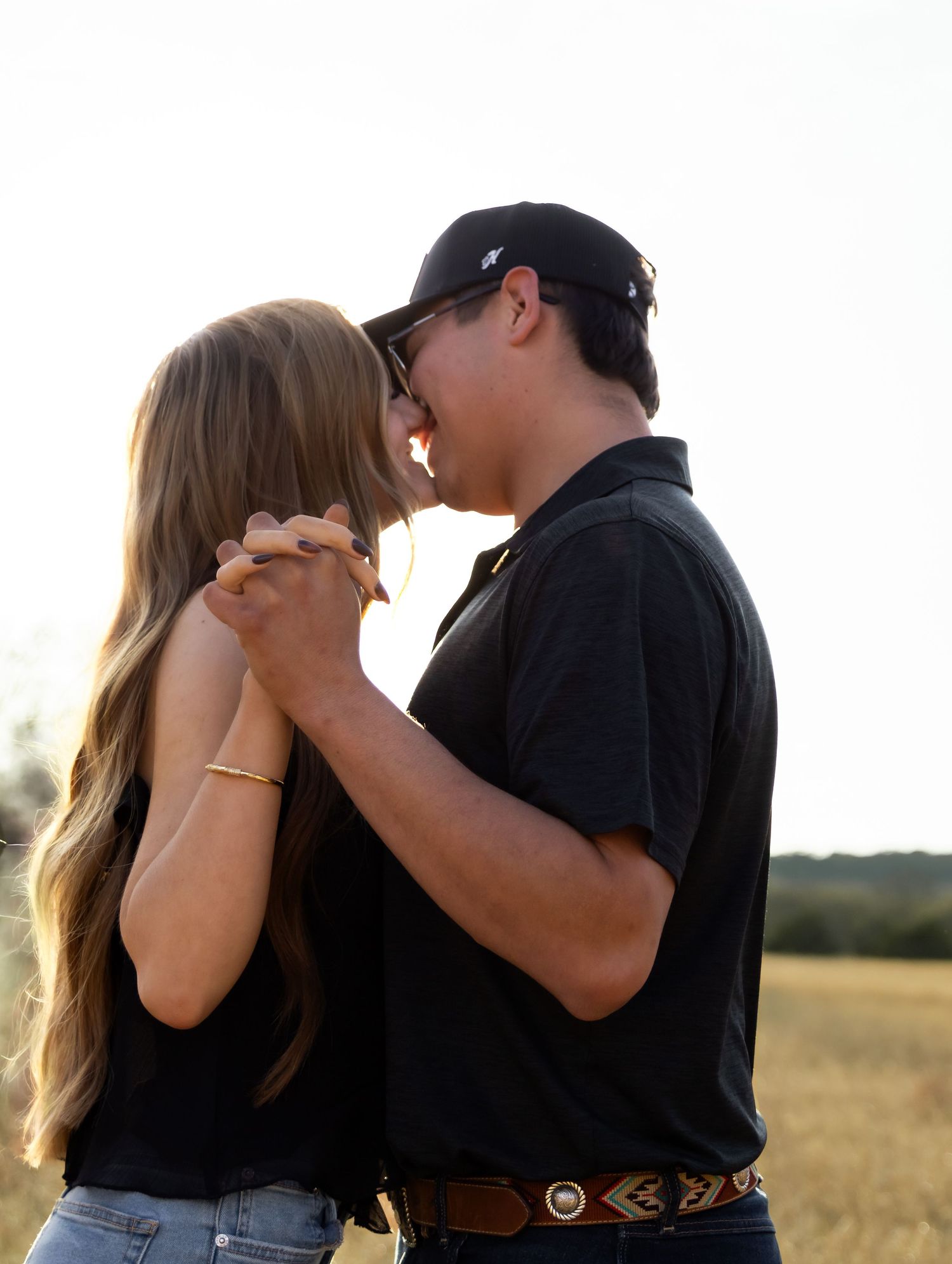 Romantic couple nearly kissing at golden hour in open field, both dressed in black.