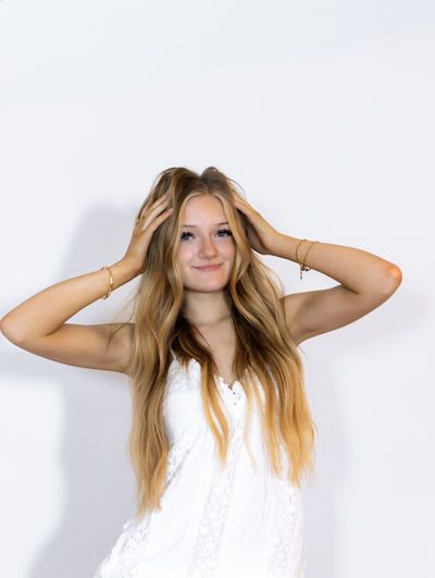 Young blonde woman in white lace dress posing with hands in hair against white wall.