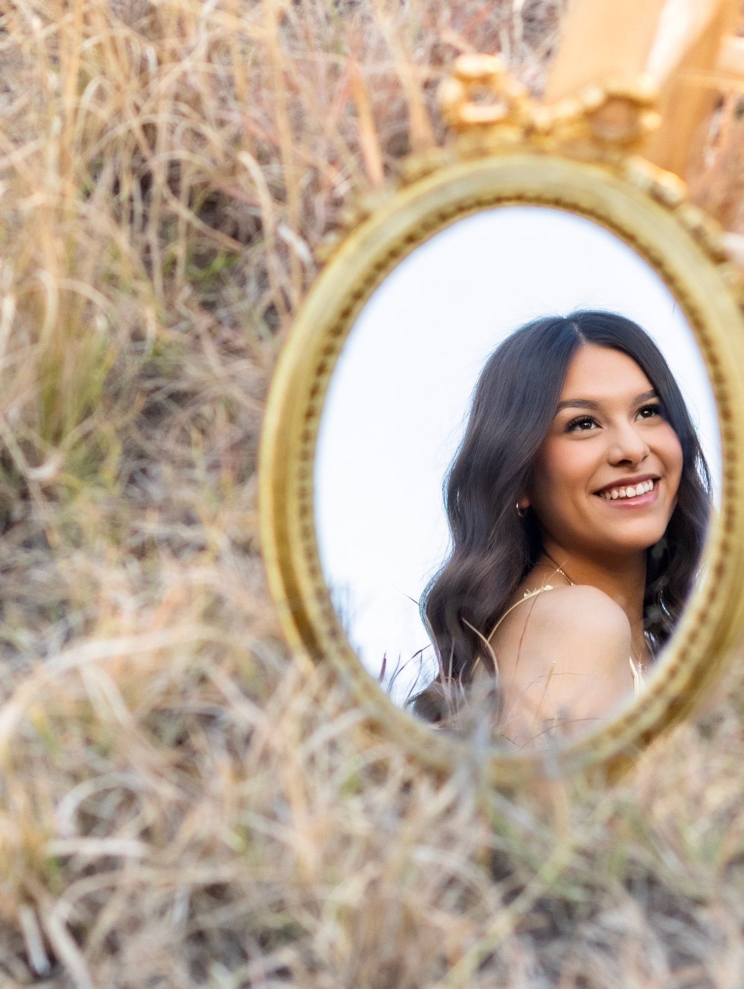 Young woman smiling reflected in ornate gold oval mirror placed in dry grass field.