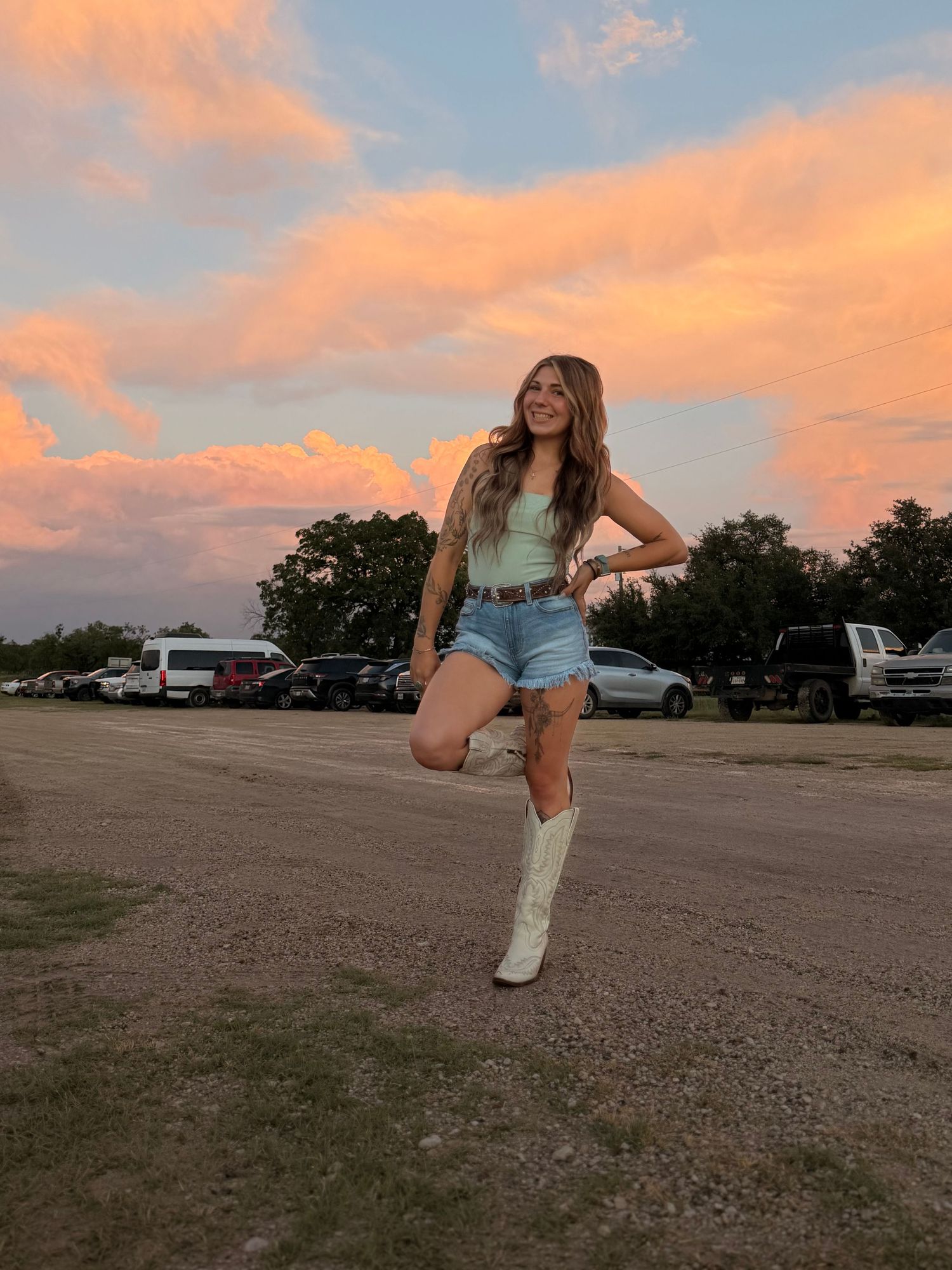 Woman in mint top, denim shorts, and white cowboy boots smiling at vibrant sunset.