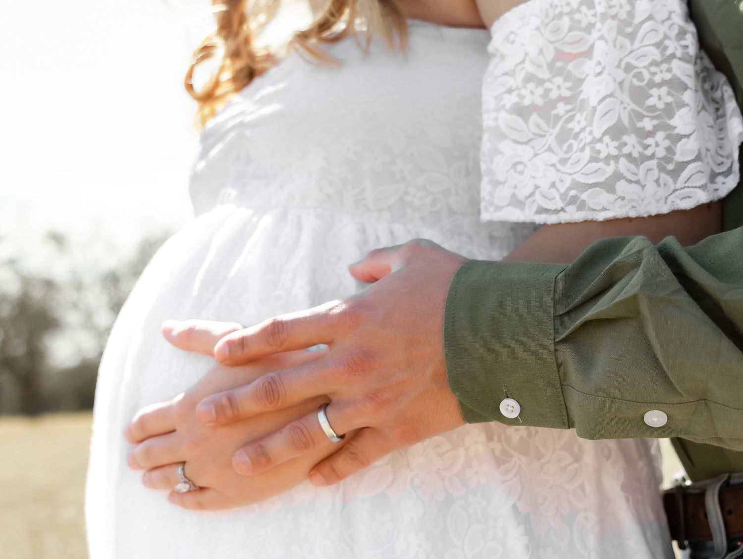 Close-up of couple embracing pregnant woman's belly outdoors, both wearing wedding rings.