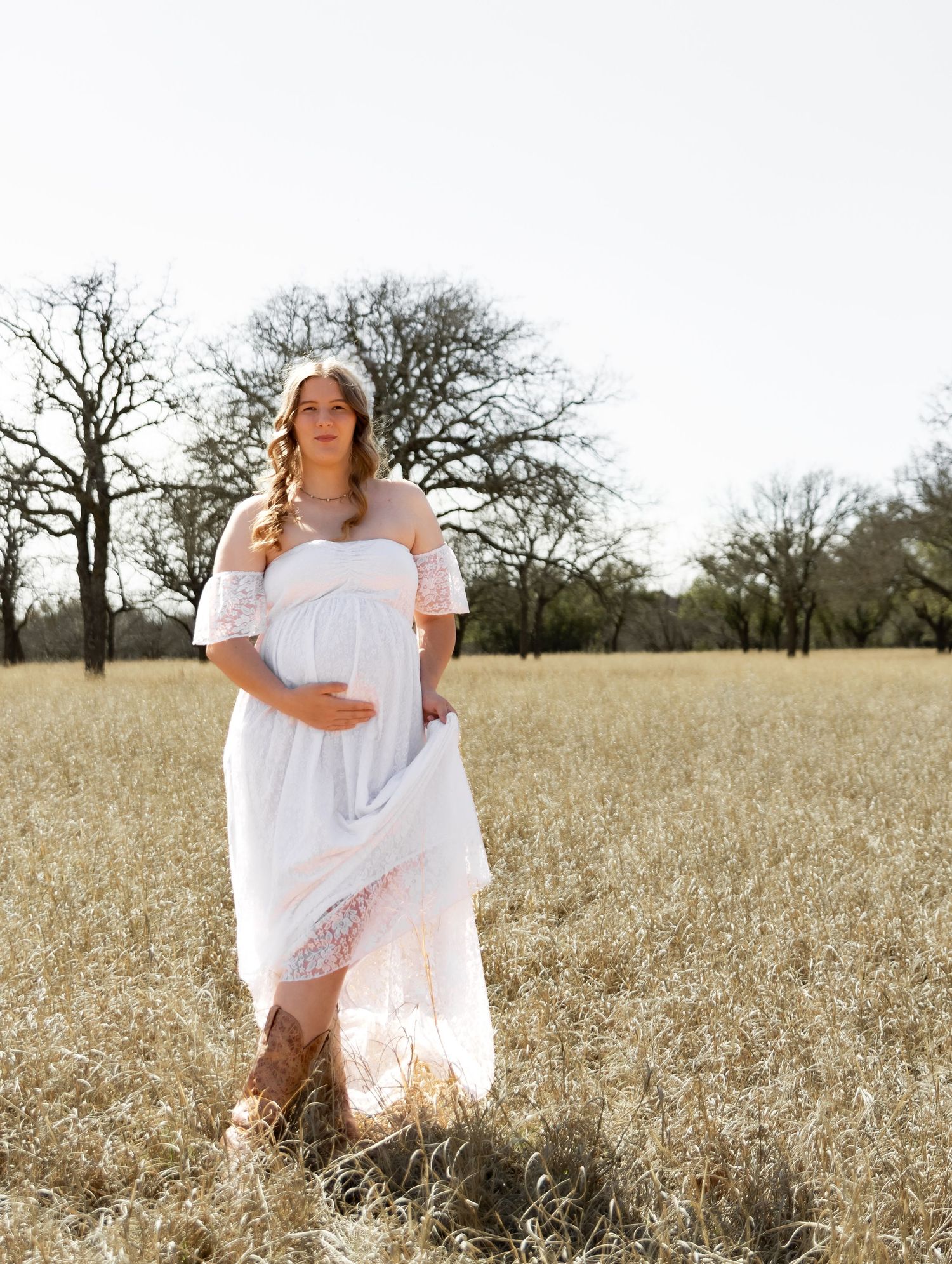 Expecting mother in white lace maternity dress holding baby bump in sunlit autumn field.