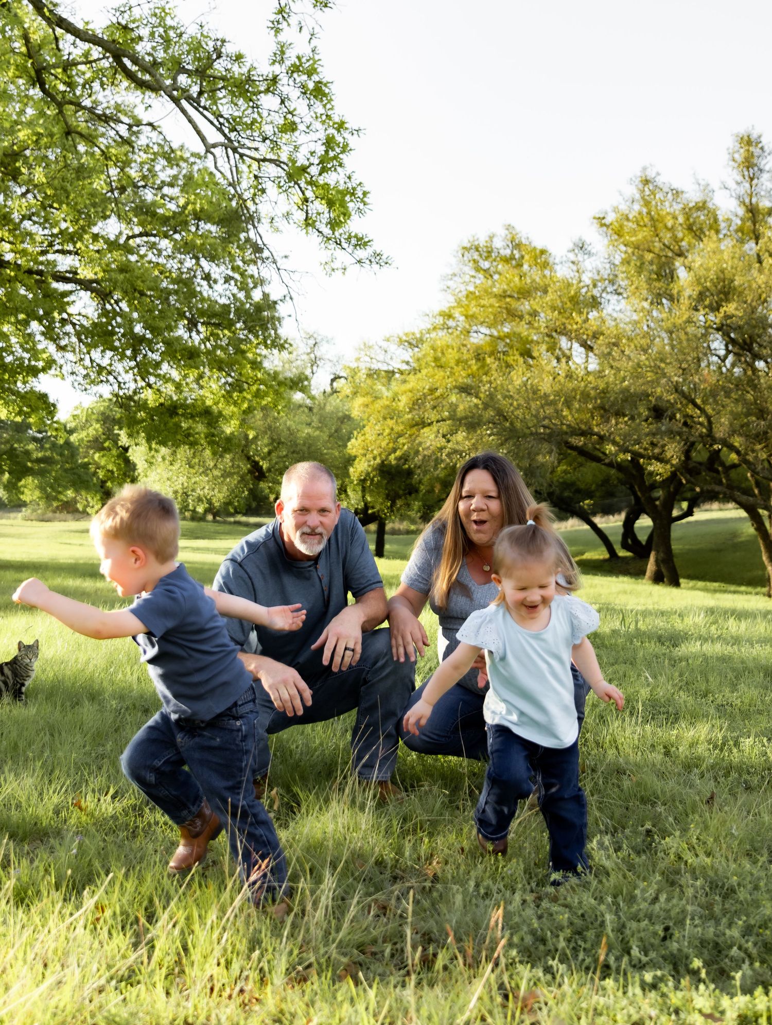 Joyful family laughing as two young children run toward the camera in a sunny green field.