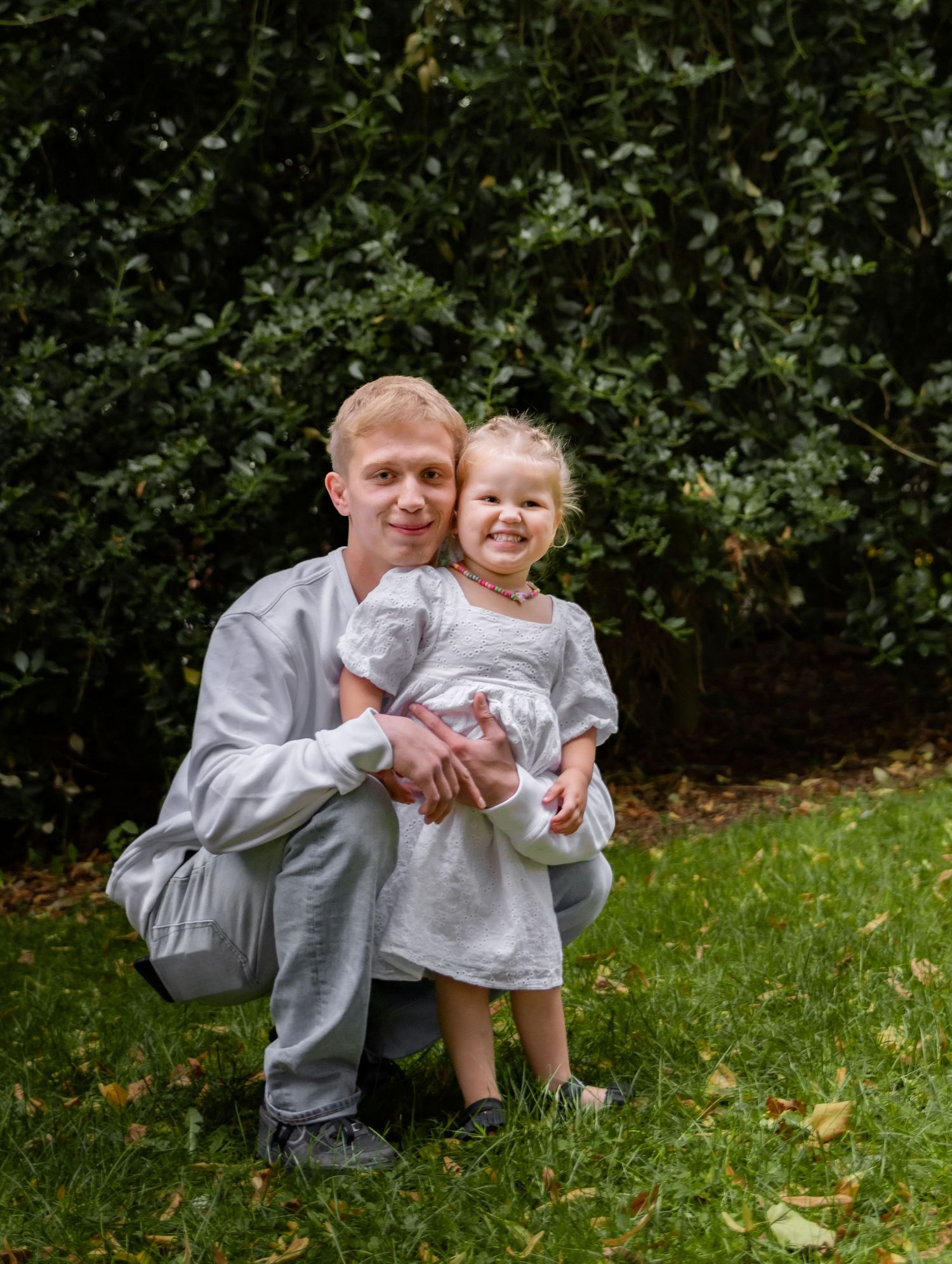 Man in gray crouching with smiling toddler girl in white dress on green lawn with bushes background.
