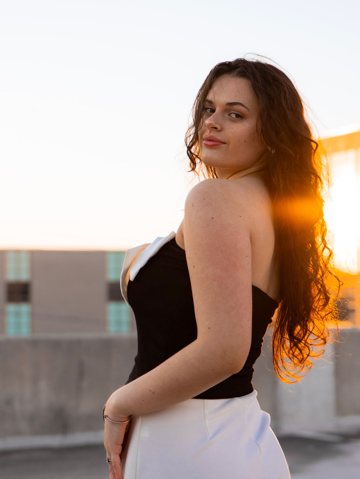 Brunette woman in black strapless top and white pants posing at sunset on rooftop.