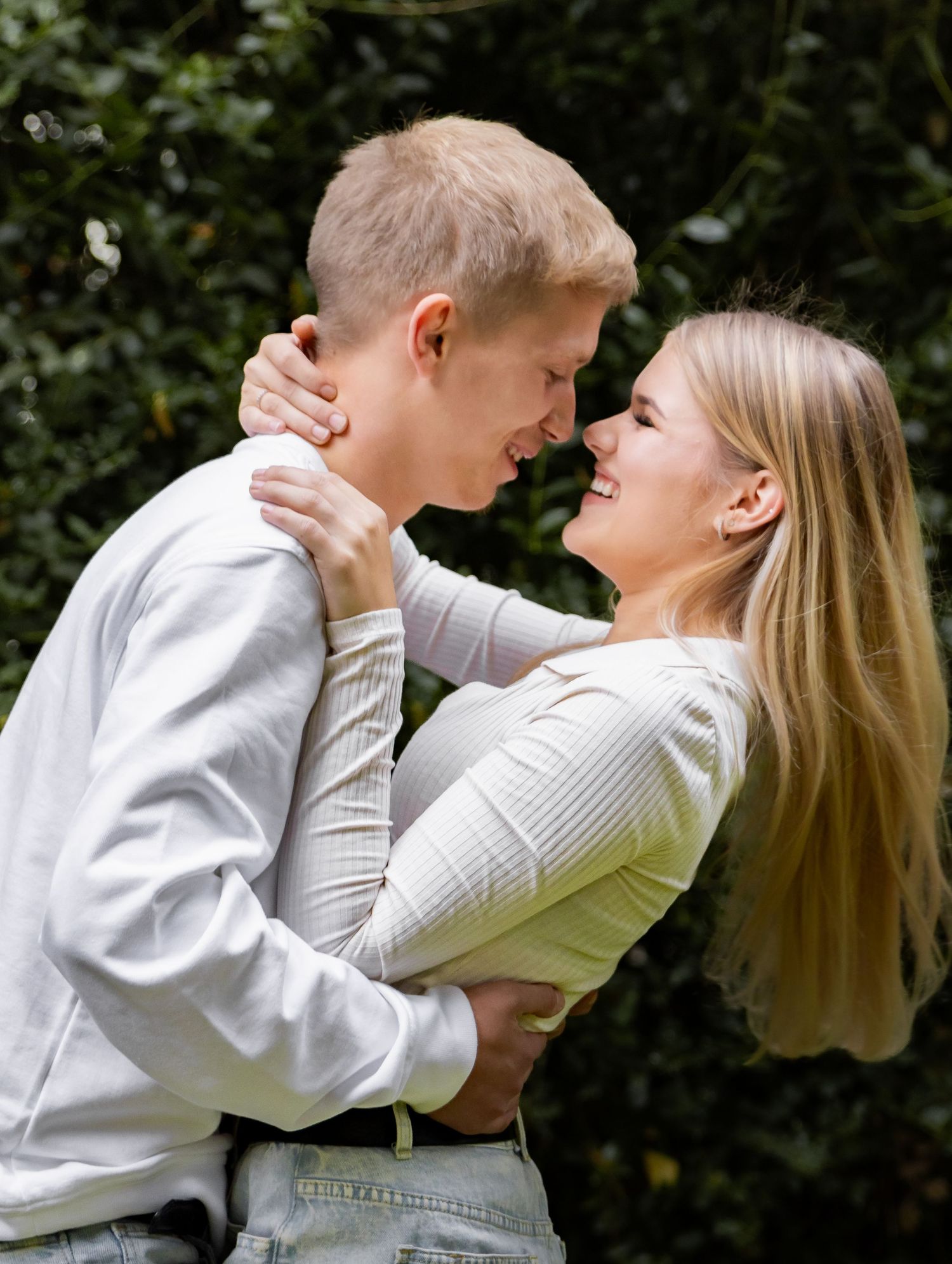 Happy young couple embracing and smiling face to face in front of green garden backdrop.