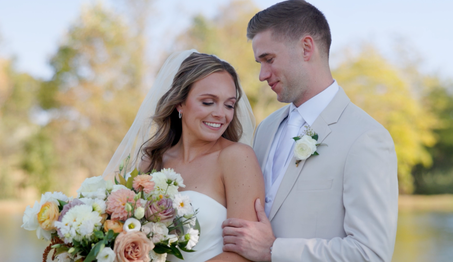 Couple in wedding attire posing outdoors during autumn with golden foliage in background.