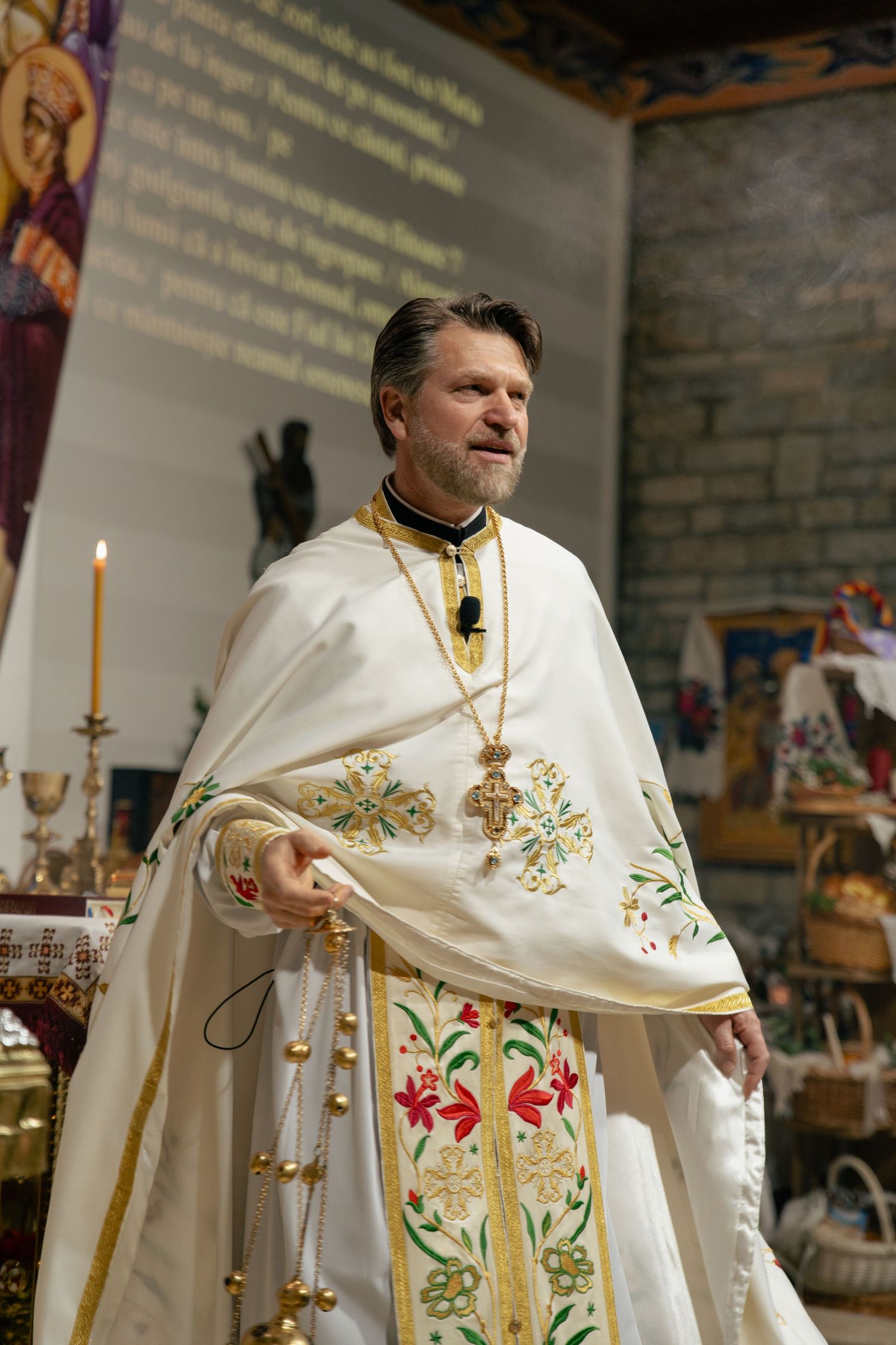 Orthodox priest in white embroidered vestments holding a censer during a religious ceremony inside a church.