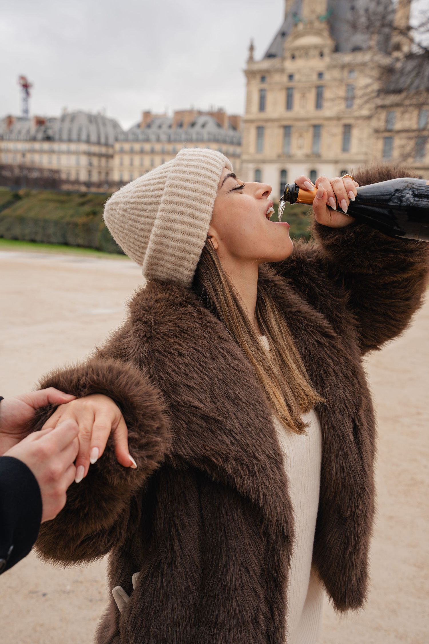 A person wearing a brown fur coat and beige beanie poses dramatically in front of a grand European palace.