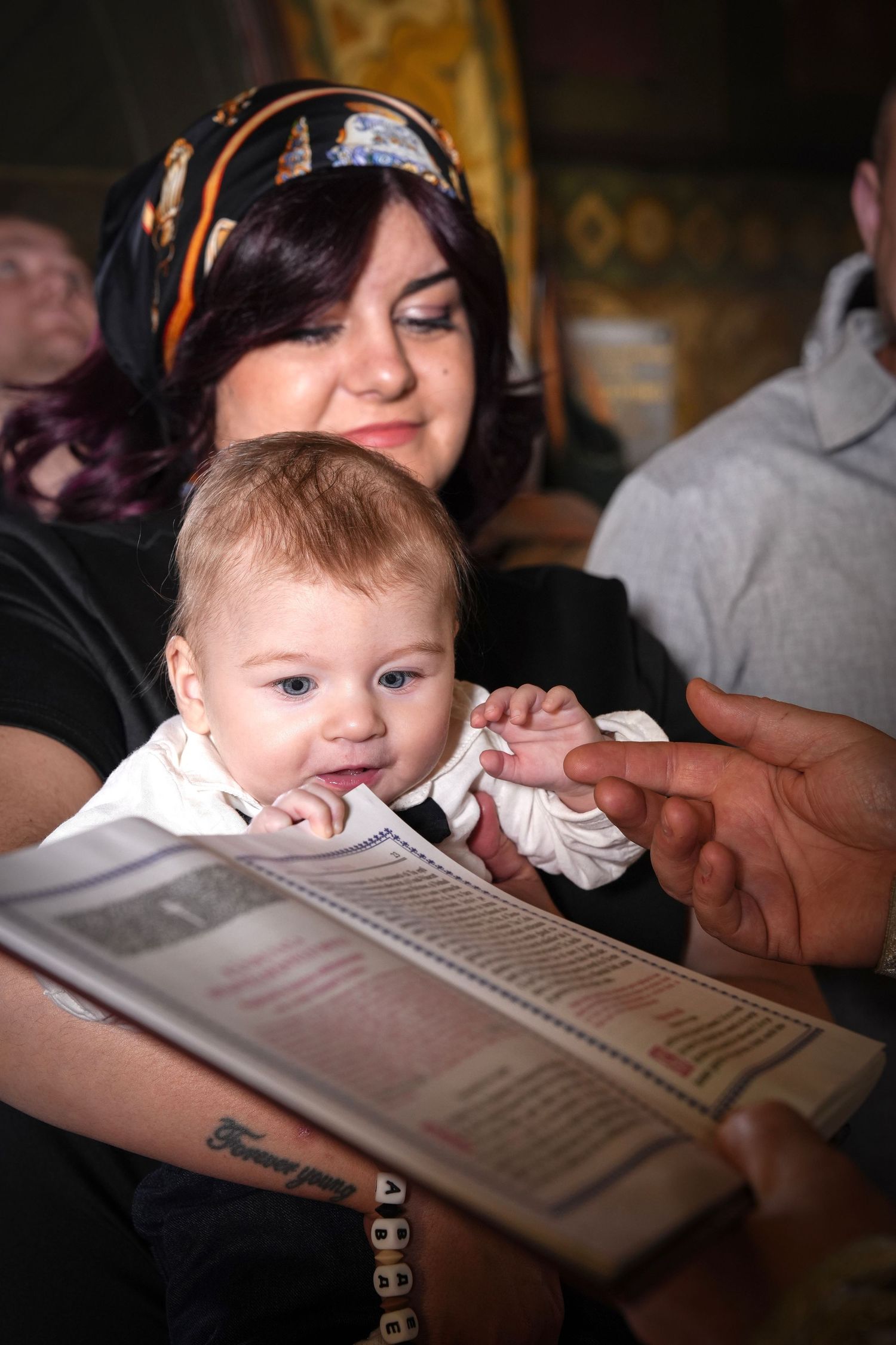 Woman in headscarf holds baby at a religious christening ceremony while an adult points at a prayer book.