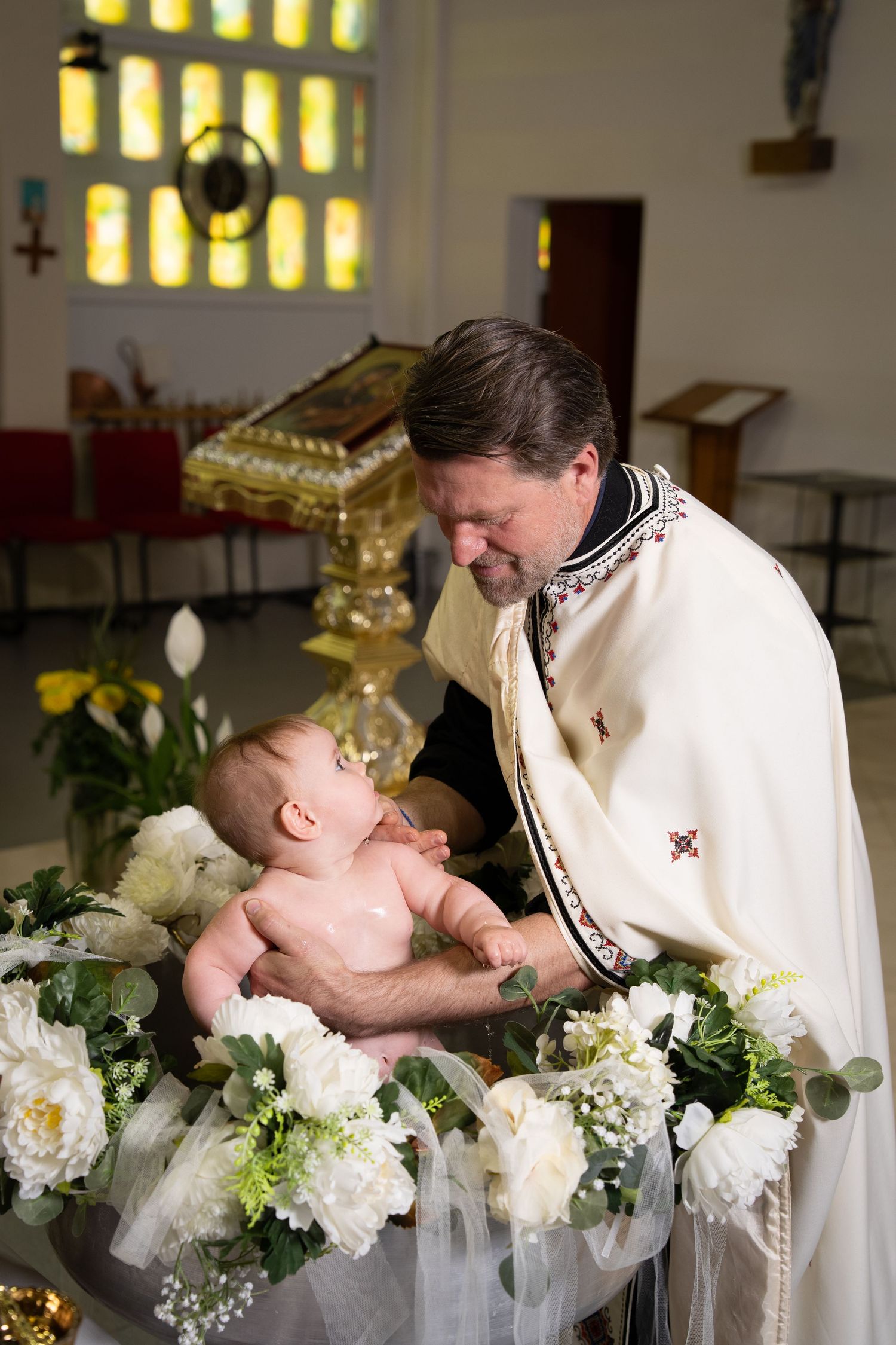 Priest in white vestments performing a baby baptism ceremony at a church font decorated with white flowers.