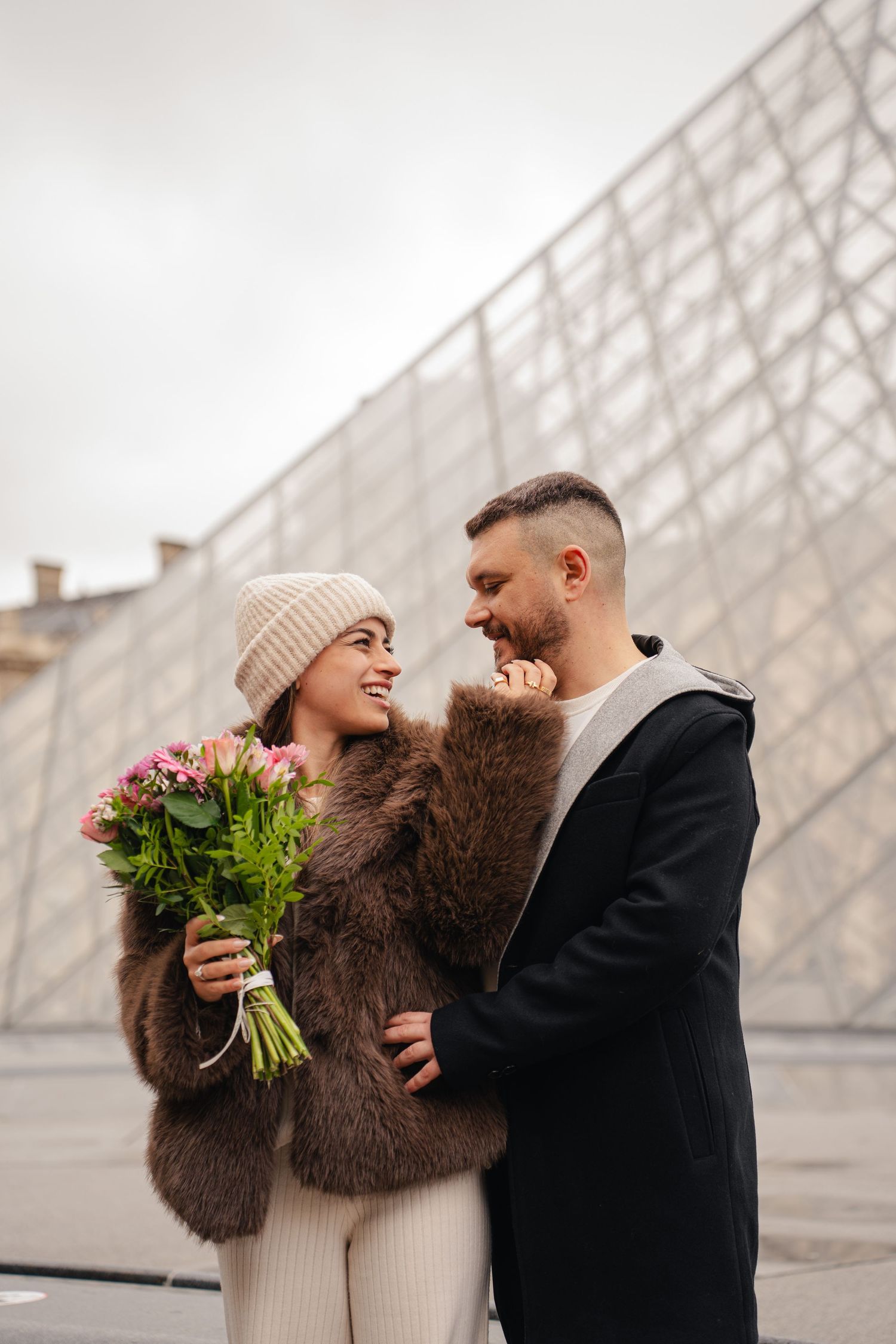 Couple shares romantic moment near geometric glass building while holding pink flowers on cloudy winter day.