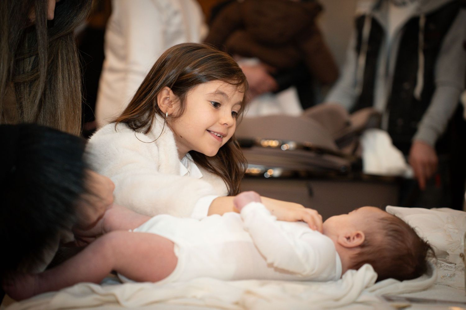 A young child in white clothing gently leans over a baby lying on white bedding.