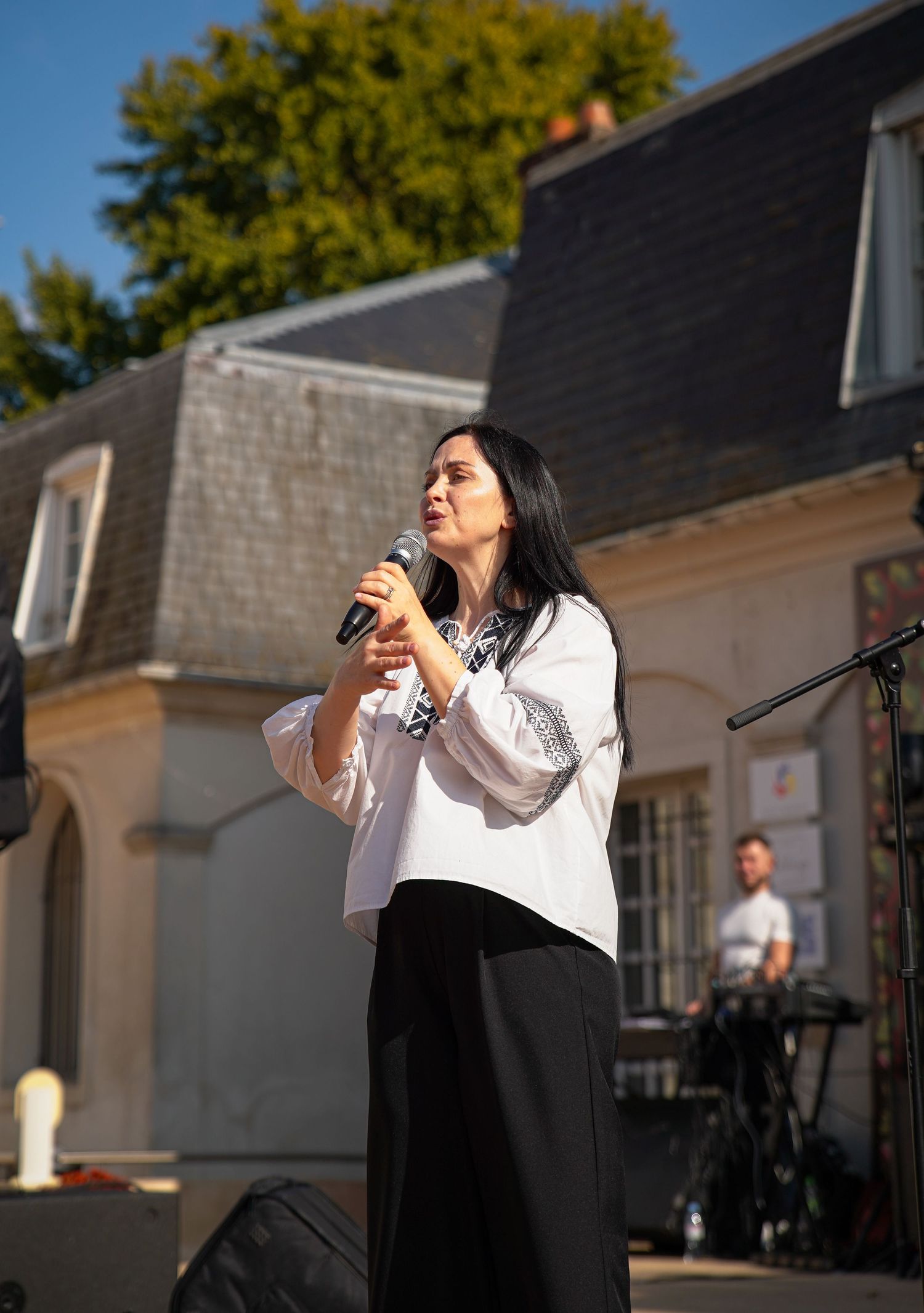 Woman singer performing outdoors holding microphone, wearing white blouse and black skirt, with historic building backdrop.