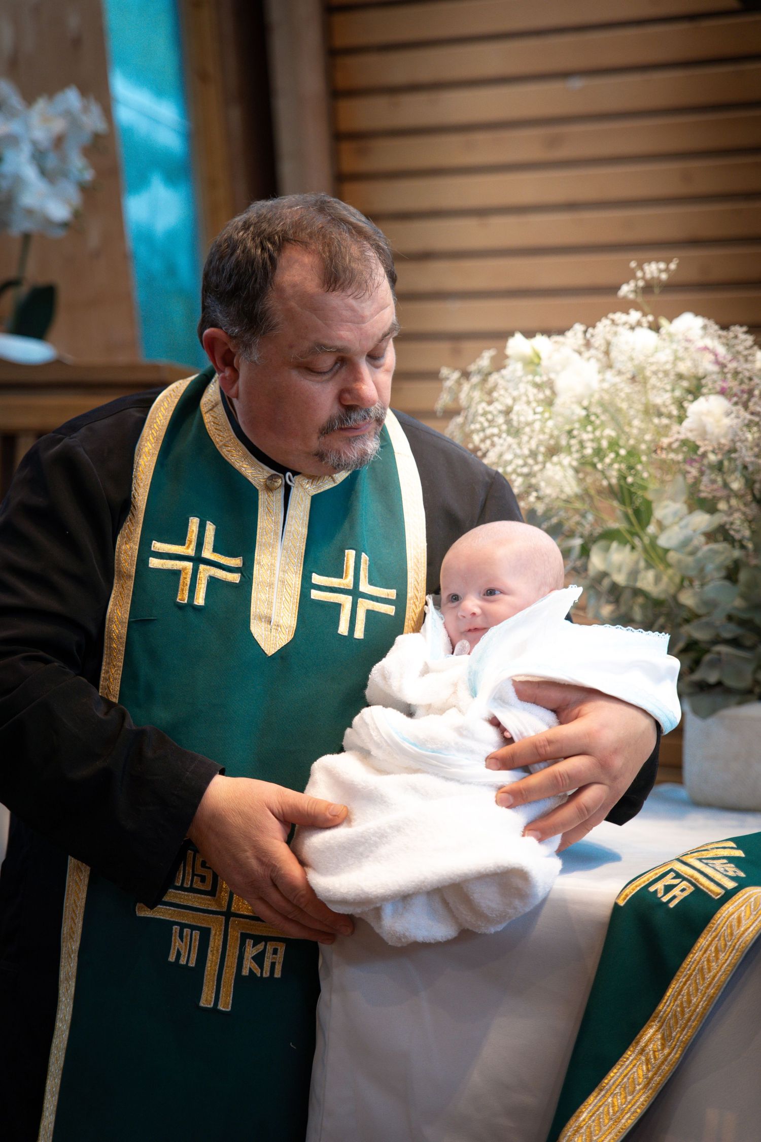 Prêtre vêtu de vêtements liturgiques verts et dorés tenant un bébé lors d’une cérémonie de baptême.