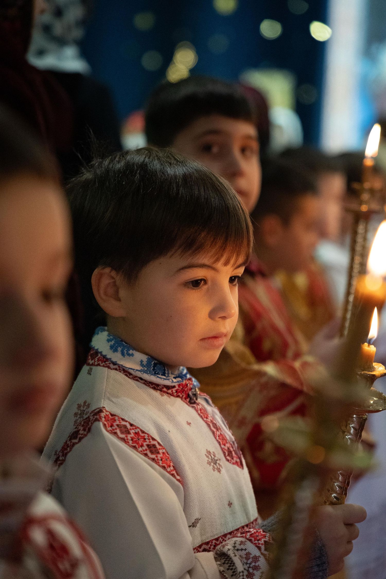 Child in traditional white embroidered outfit holding candle during religious ceremony.