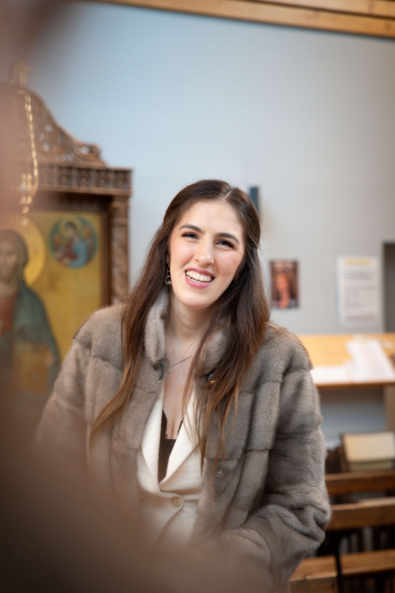 Smiling young woman in gray fur coat laughing in a church interior with religious icons in background.