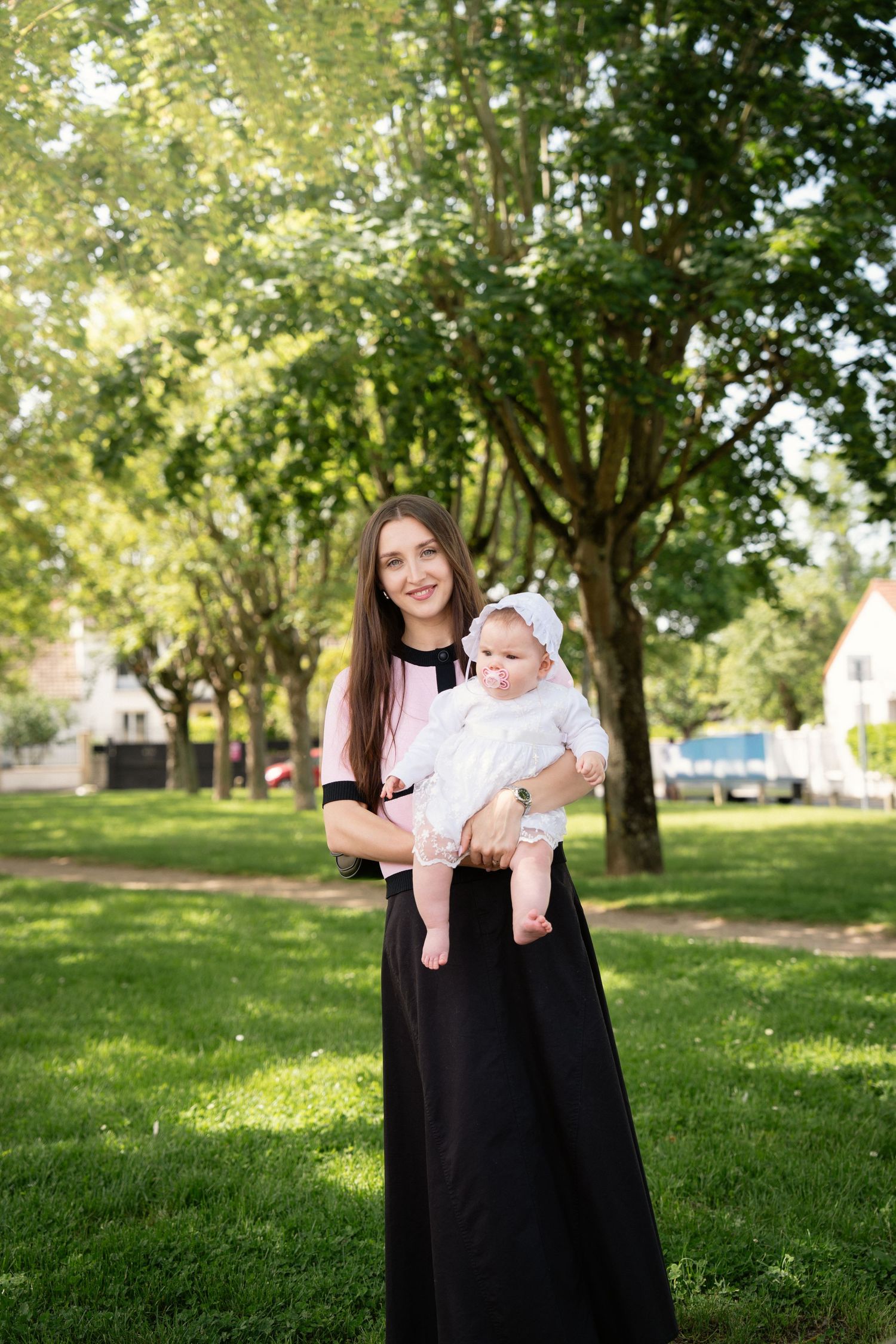 Young mother in black skirt holding smiling baby in white dress outdoors in green park on sunny day.