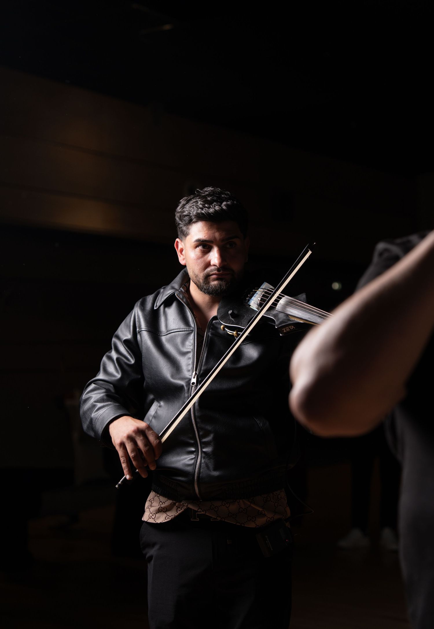 A man in a black leather jacket plays violin on a dimly lit stage with focused intensity.