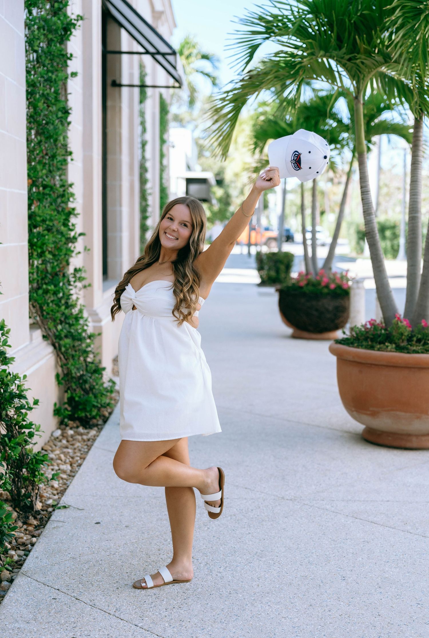 Young woman in white mini dress holding Instax camera aloft on sunny Palm Beach sidewalk.