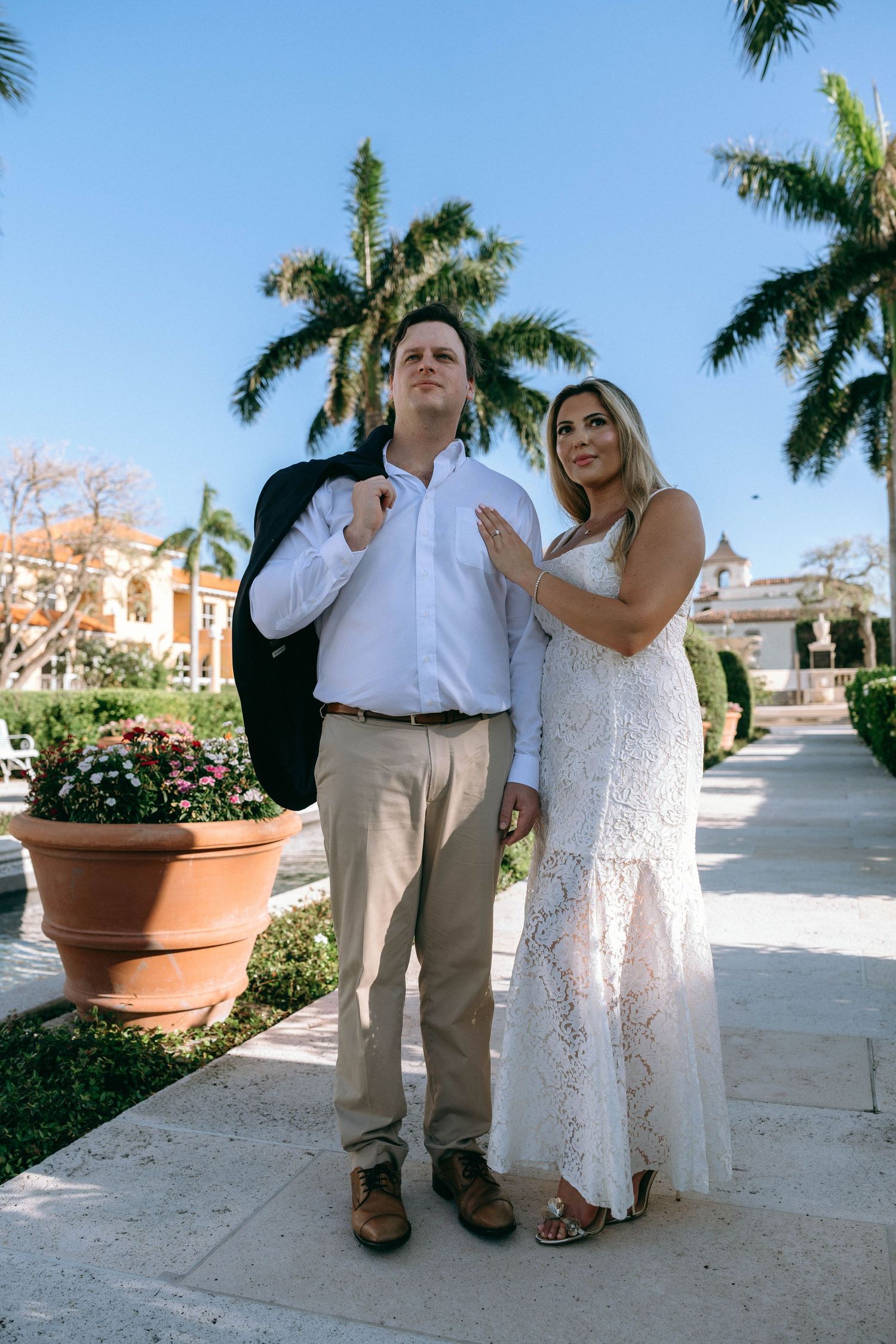 Couple posing outdoors near palm trees, woman in white lace dress and man in white shirt with blazer.