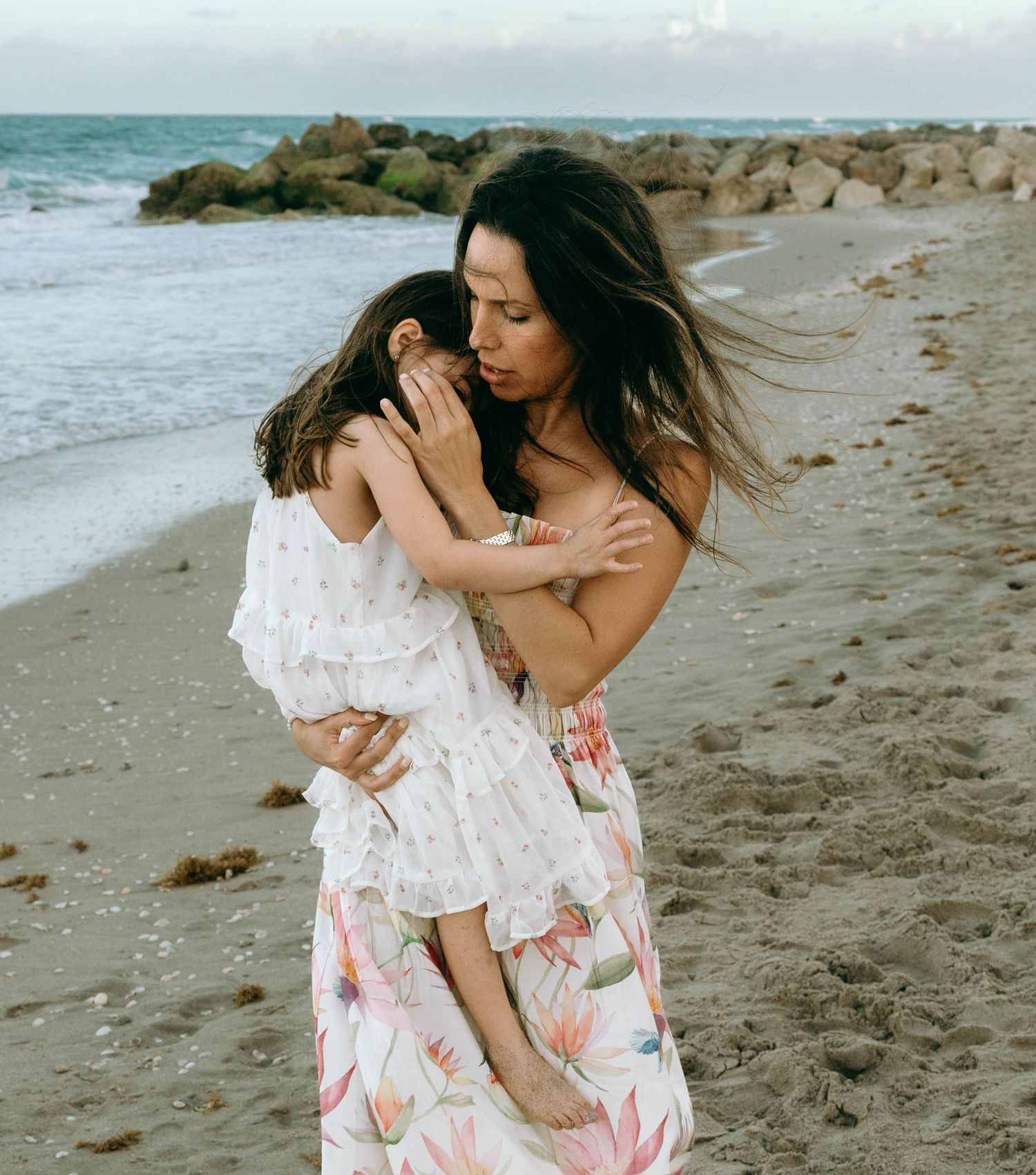 A mother tenderly holds her young daughter on a sandy beach, both wearing floral dresses near rocky shoreline. Worth Avenue, Palm Beach, FL