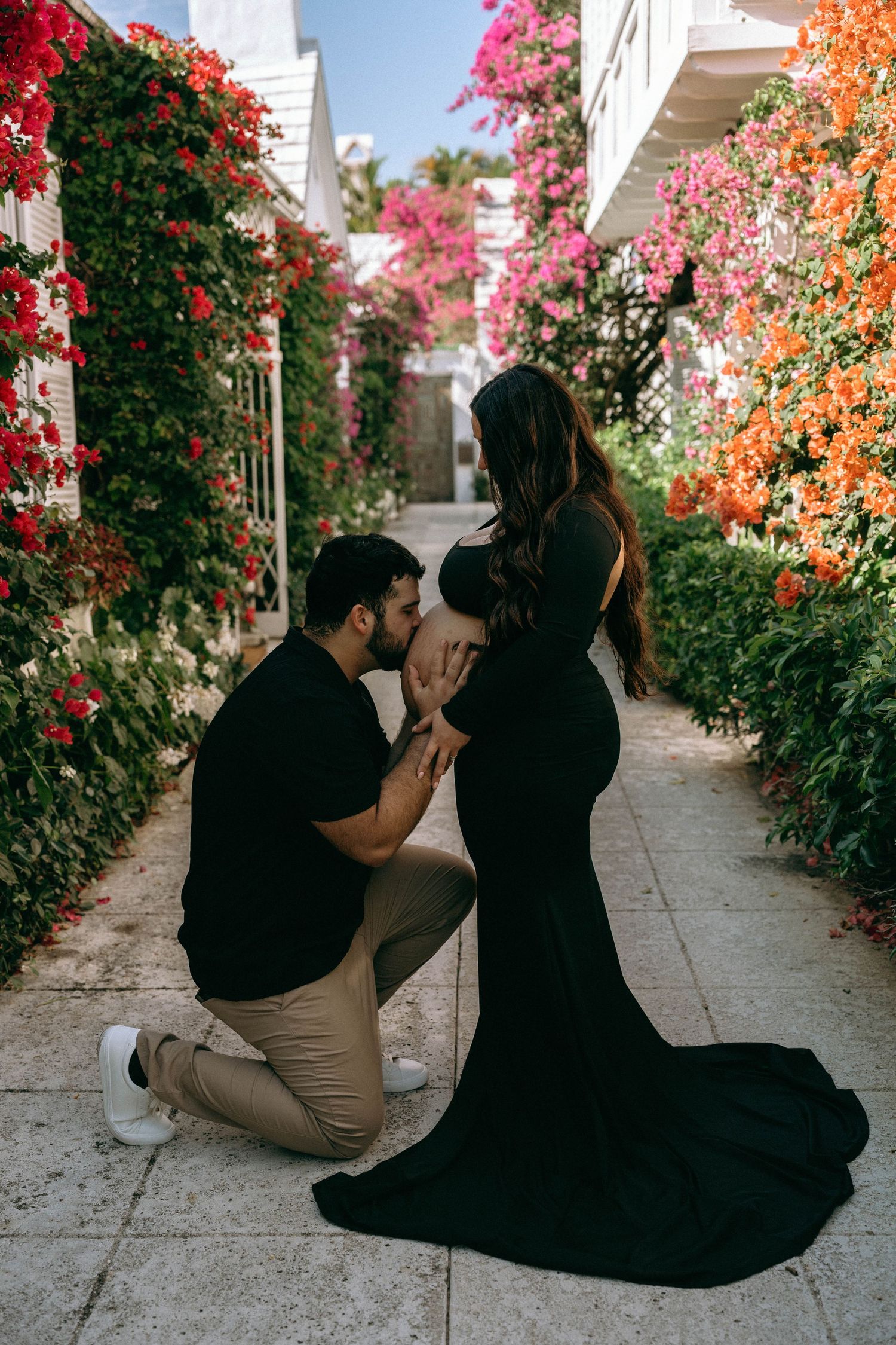Man kneeling and kissing pregnant woman's belly in a colorful flower-lined garden pathway. Worth Avenue, Palm Beach, FL