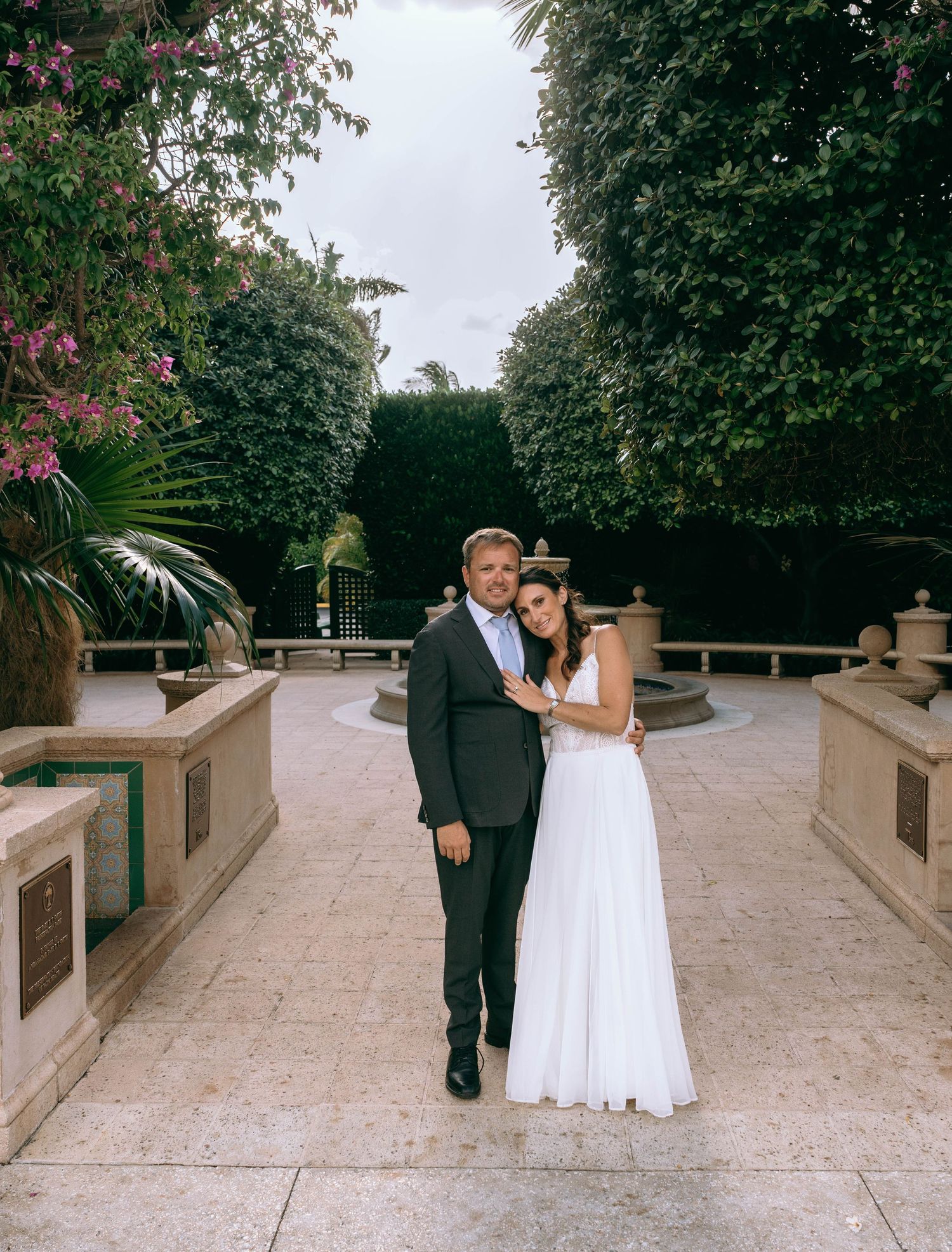 Bride and groom posing together in a lush garden courtyard, bride wearing a white gown and groom in a suit.