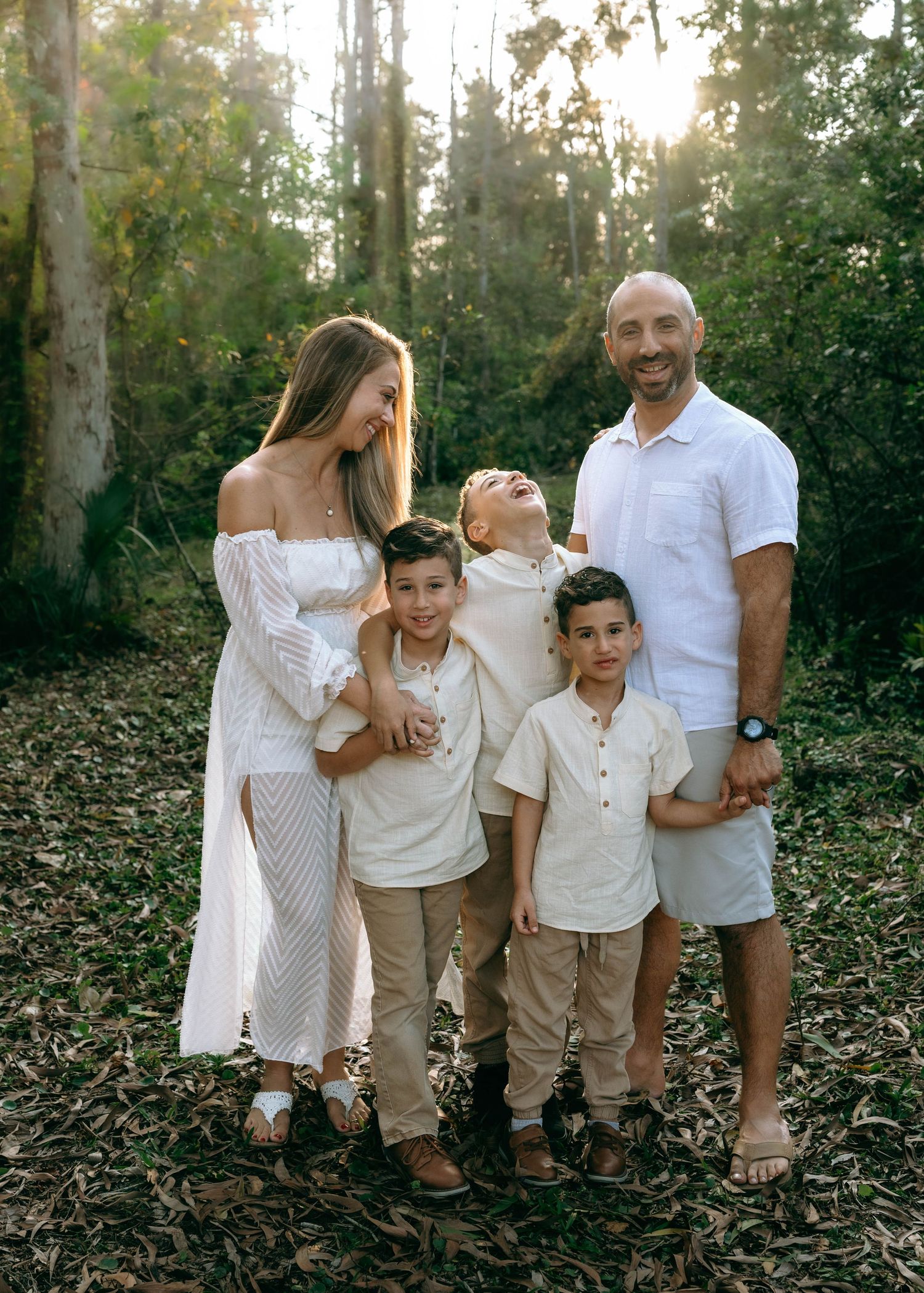 A family in coordinating white and beige outfits poses playfully together in a wooded outdoor setting.