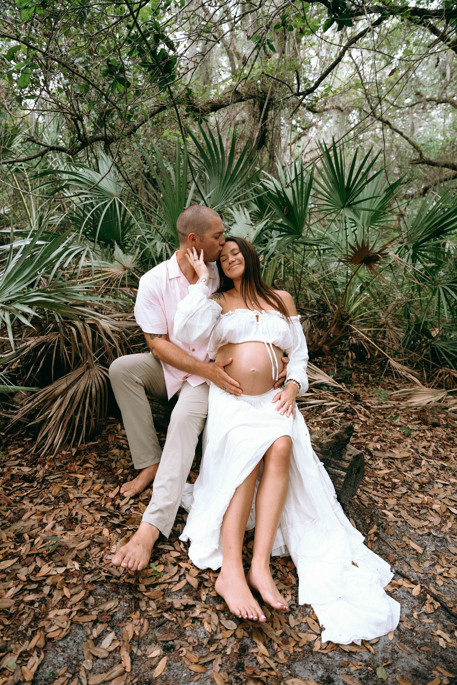Romantic couple in white clothing embrace on fallen leaves among palm trees in a natural outdoor setting.