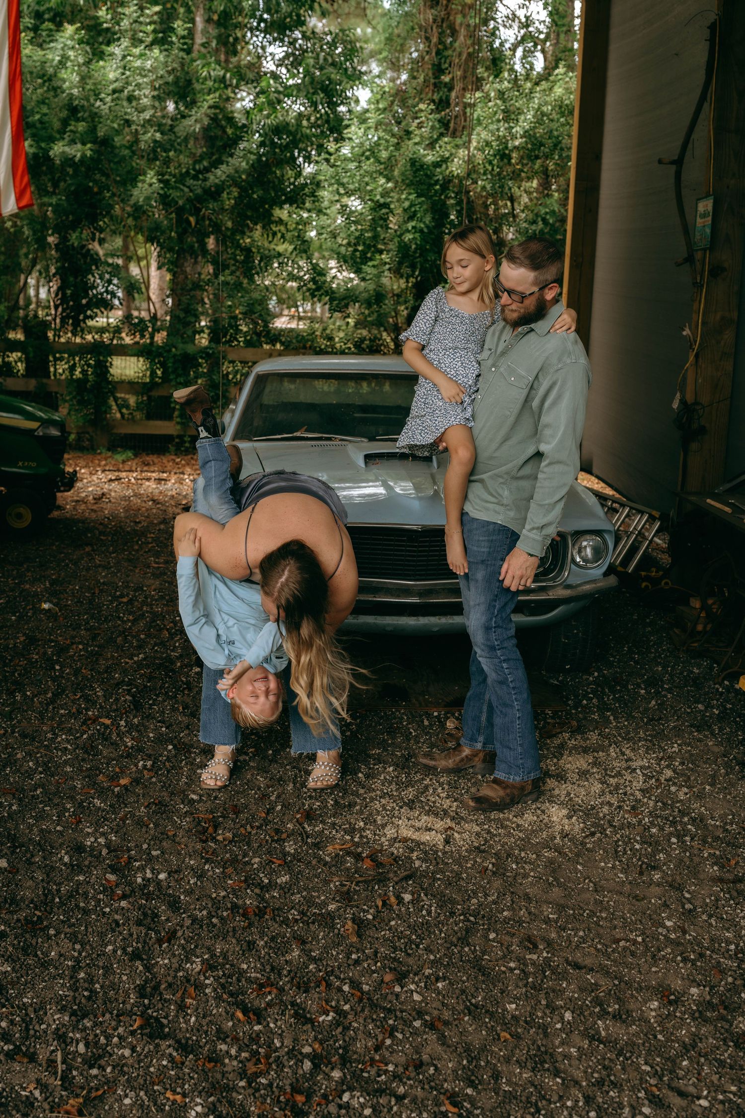 Family posing playfully in front of a vintage car, with a man holding a child and a woman bending over.