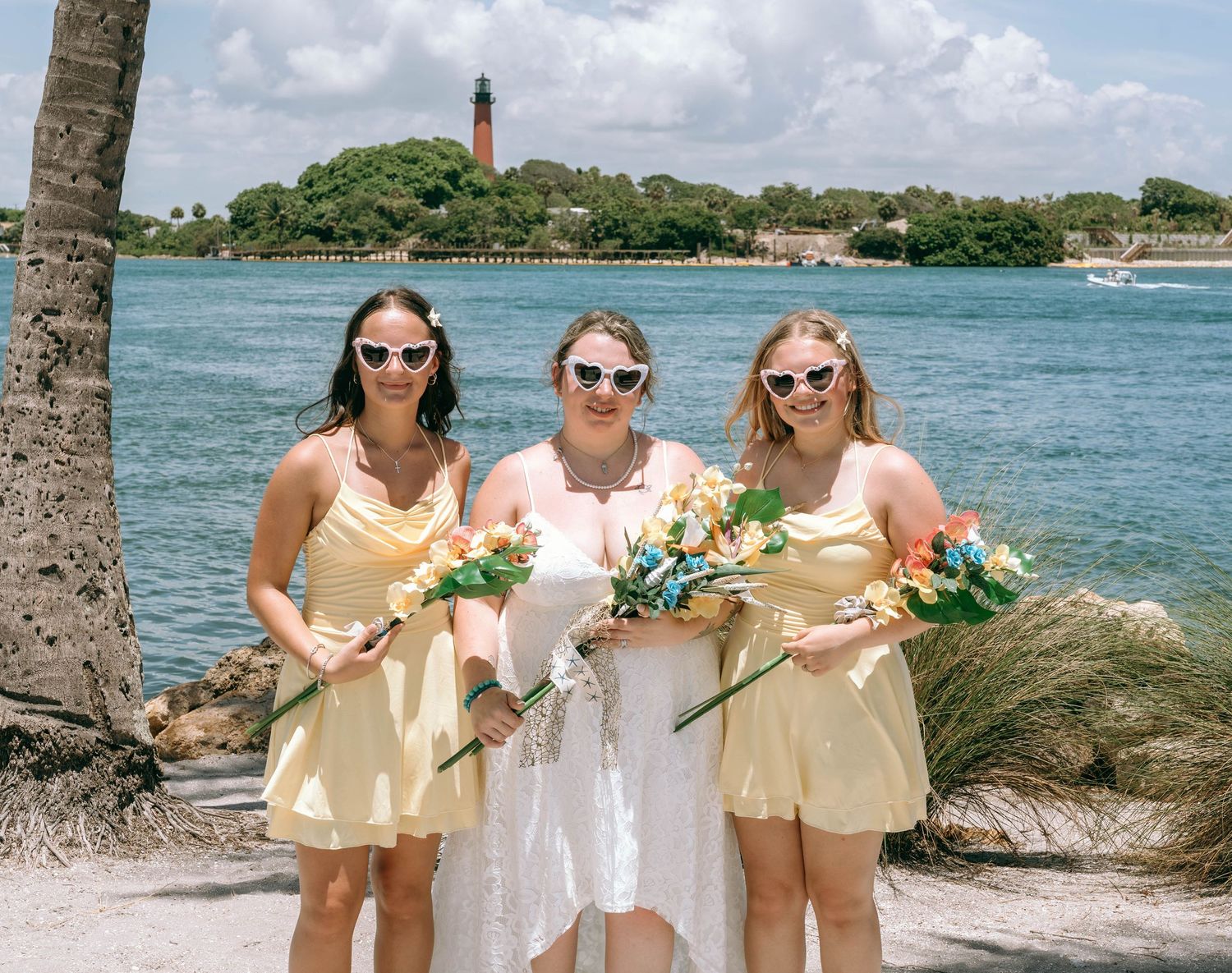 Bride with two bridesmaids in yellow dresses holding bouquets on a beach with a lighthouse in the background.