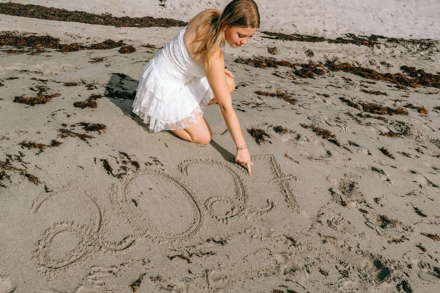 A young woman in a white dress kneels on a sandy beach, writing numbers in the sand with her finger.