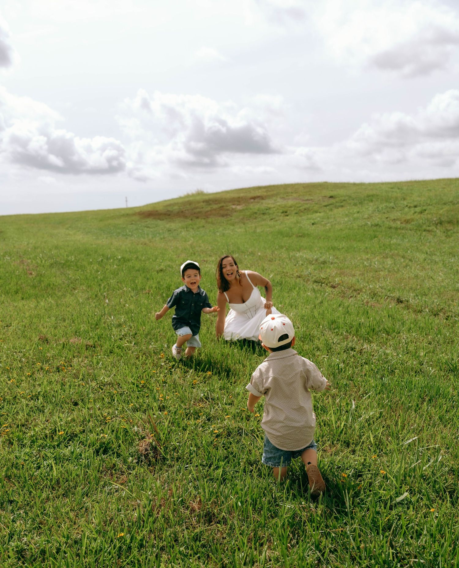 Mother and two young children playing and running on a lush green grassy hill.