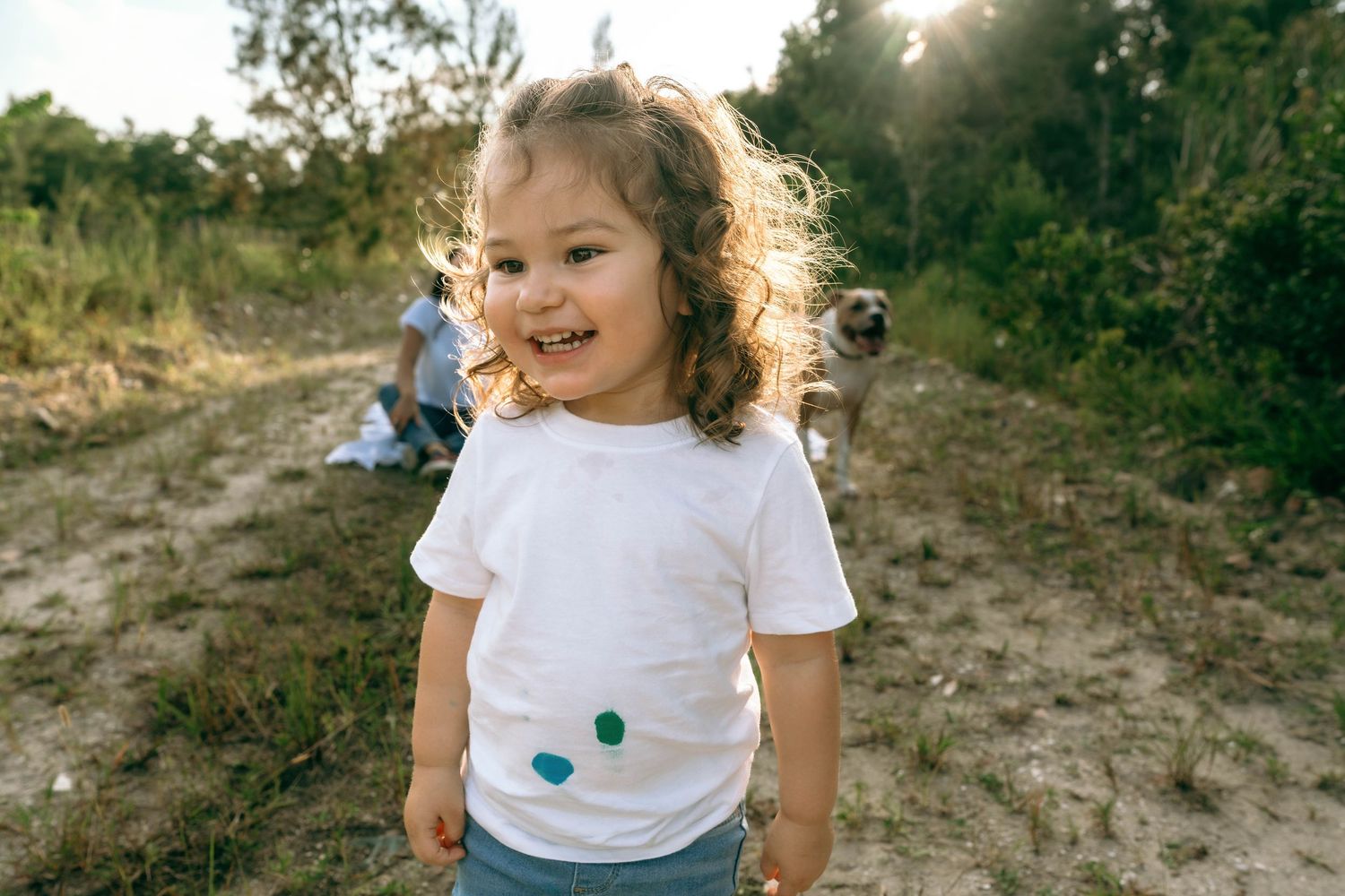 Smiling toddler girl with curly hair wearing a white t-shirt outdoors in a sunny forest clearing.