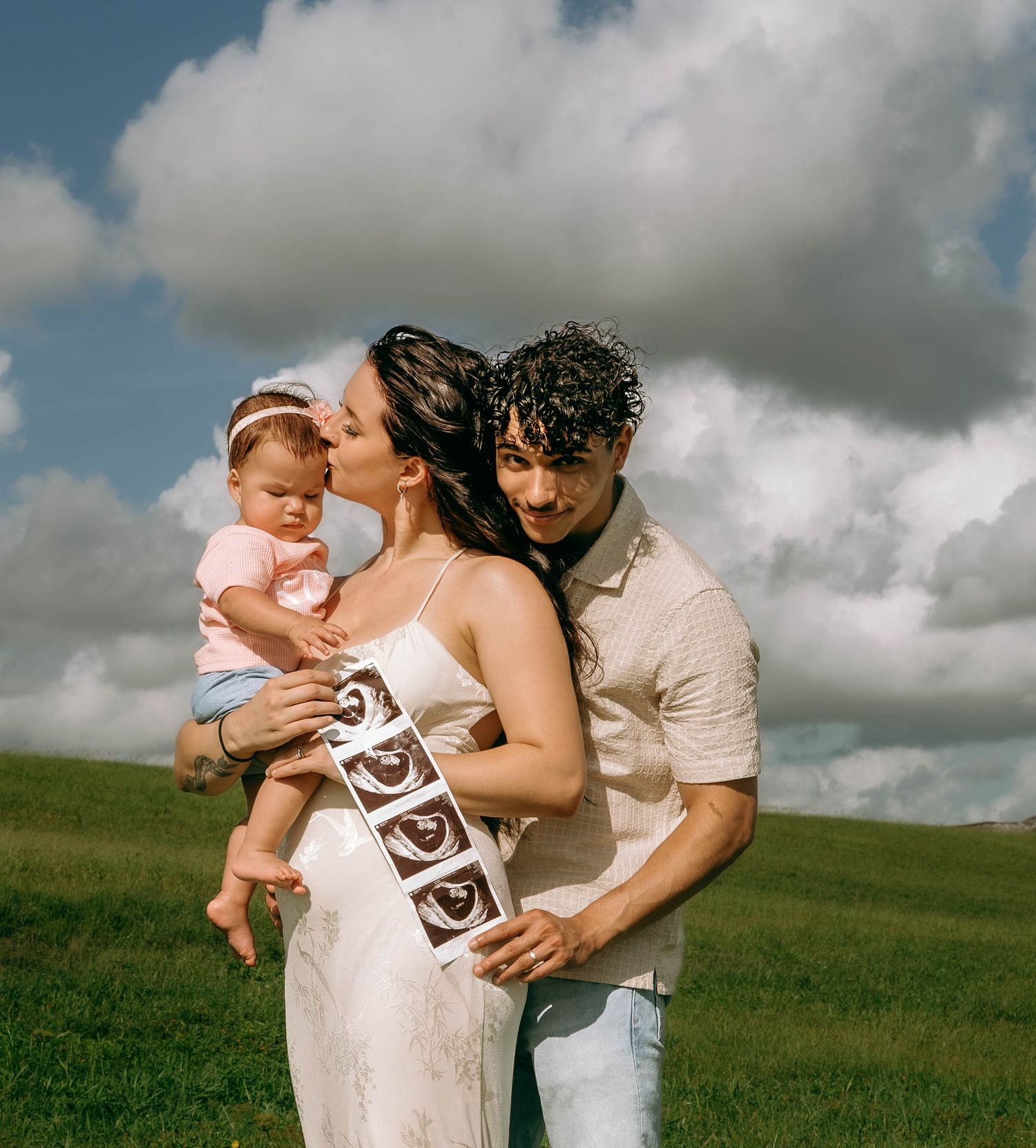 Happy family on green hill, pregnant woman kissing baby girl while man holds ultrasound strip photos.