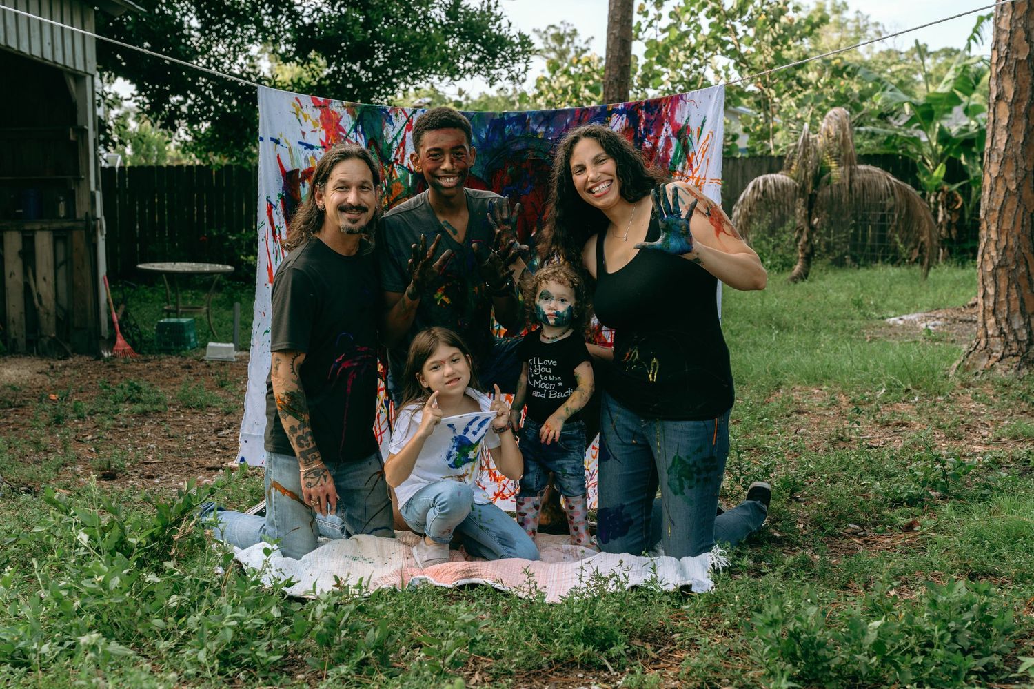 Family of four posing outdoors on a blanket with colorful painted backdrop in a backyard setting.