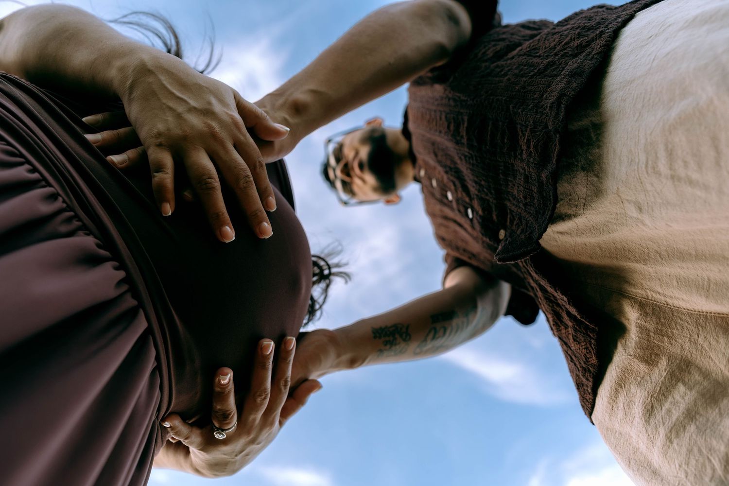 Two people tenderly cradling a pregnant belly outdoors against a blue sky, celebrating new life and motherhood.