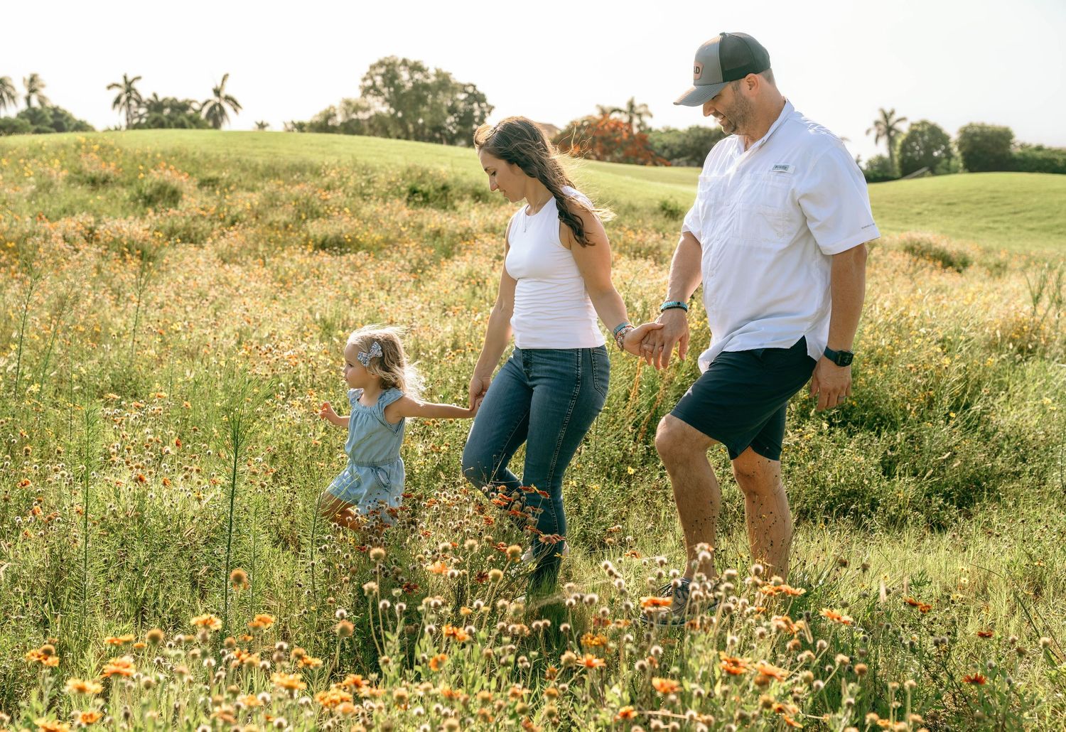 Family of three walking through a sunny wildflower meadow, parents holding child's hands on a warm day.