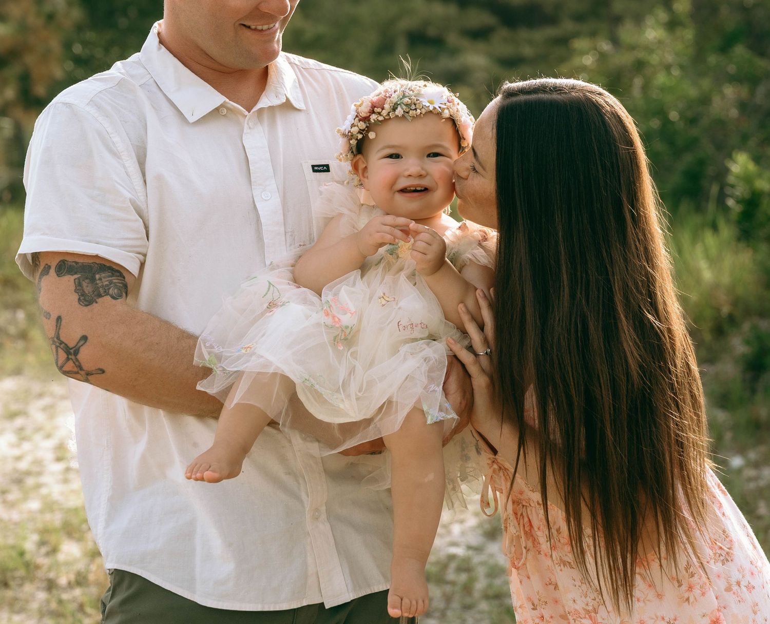 A happy family with tattooed father and dark-haired mother kissing smiling baby girl in floral dress outdoors.