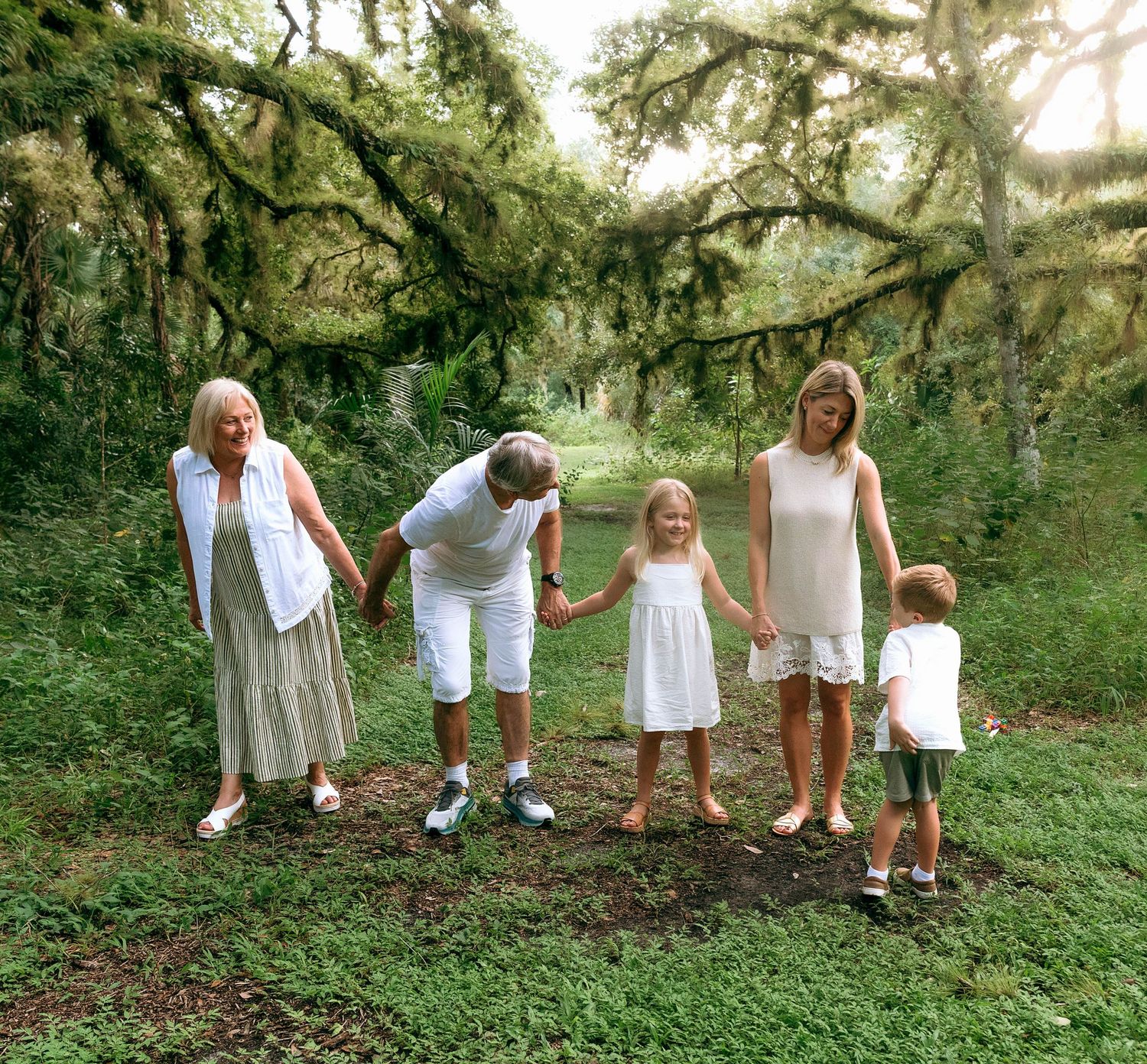 Multi-generational family holds hands in lush green forest with moss-covered oak trees at Riverbend Park in Jupiter, FL..