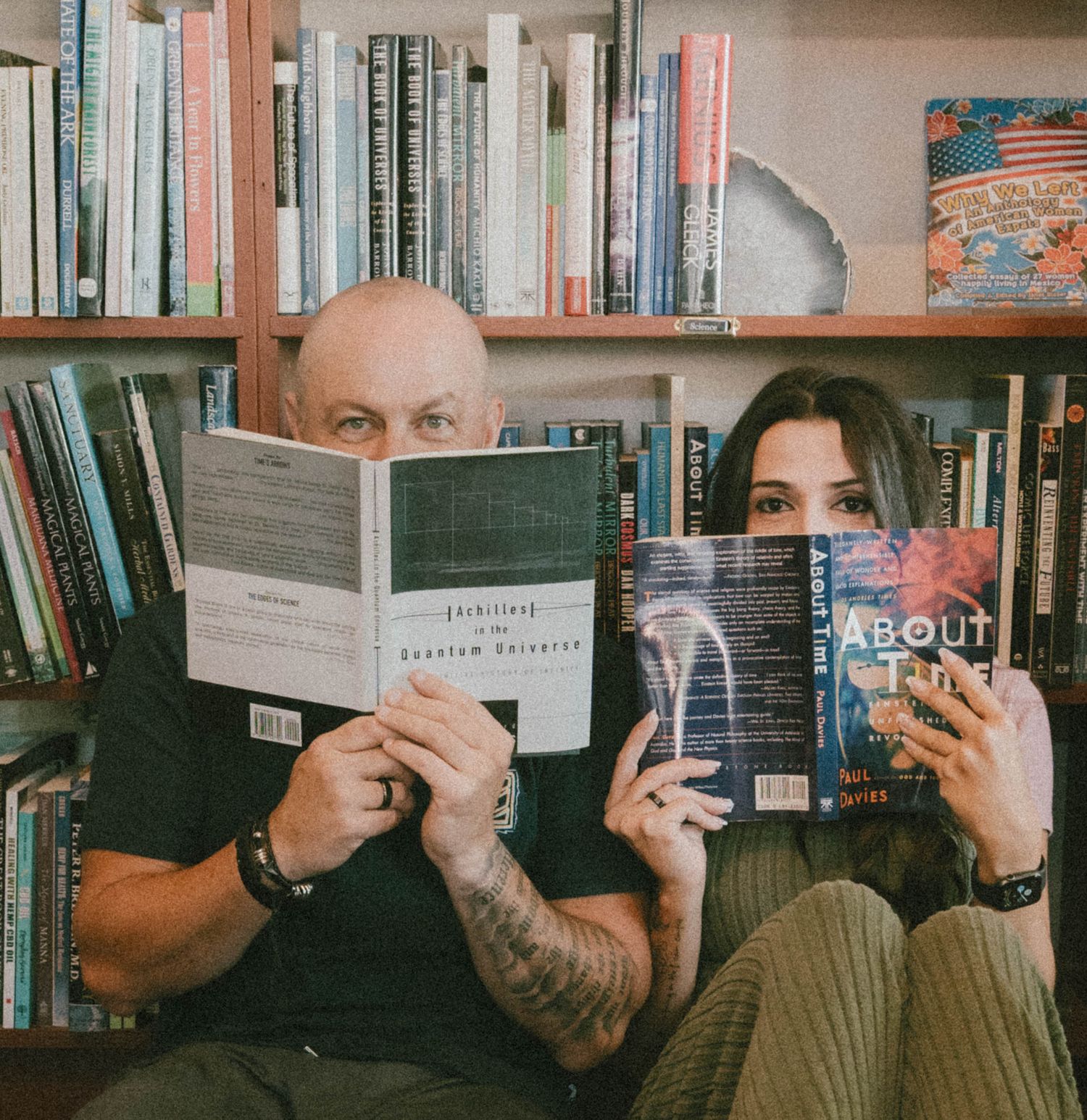 Two people reading books in front of a bookshelf, one holding About Time, in a cozy library setting.