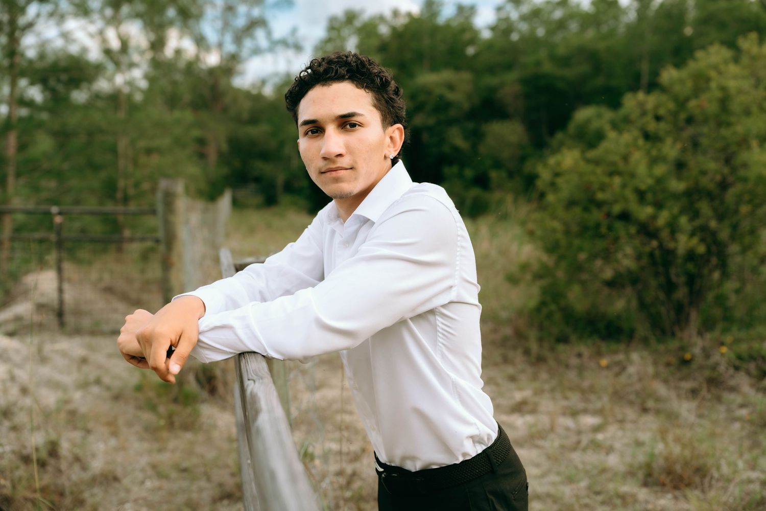 Young man in white dress shirt leaning on a fence post in a rural outdoor setting with green trees. Senior Photos at Colt Farms in Boynton