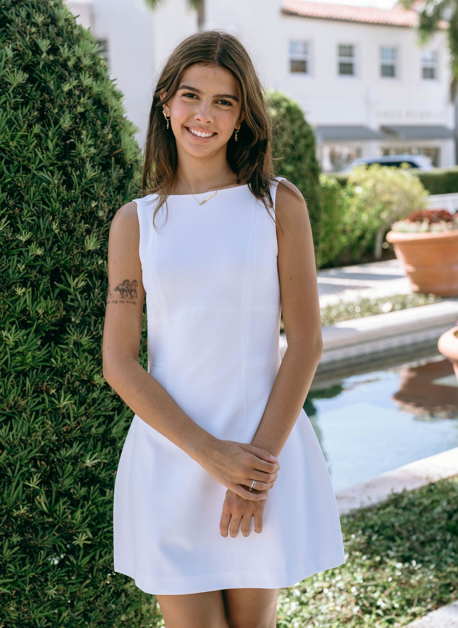 Smiling young woman in white sleeveless mini dress posing near fountain in sunny garden on Worth Avenue Palm beach.