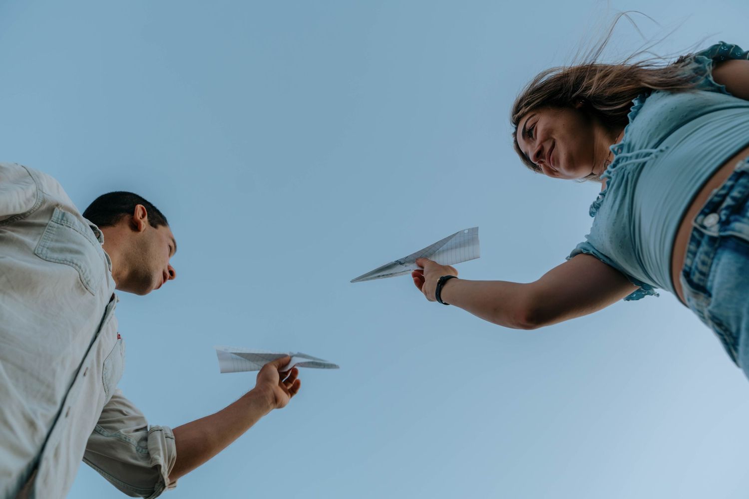 Two young people holding paper airplanes facing each other against a clear blue sky, viewed from below.