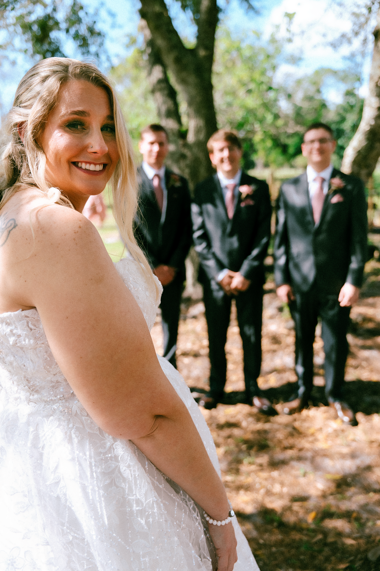 Smiling bride in white lace gown with three groomsmen in dark suits standing beneath trees at outdoor wedding.